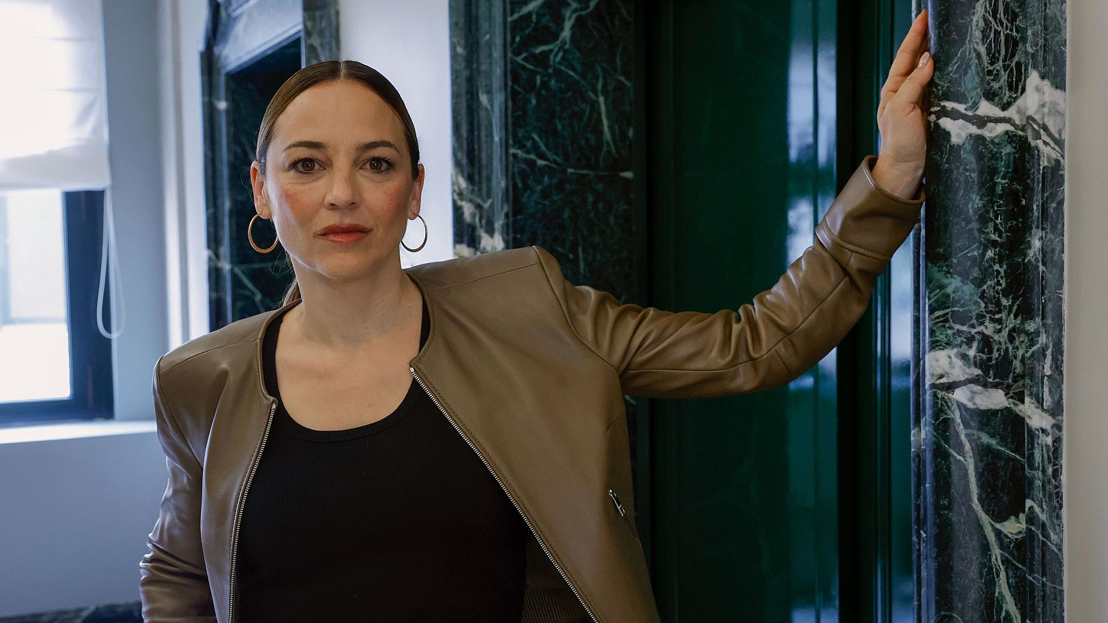 Retrato de Leonor Watling con chaqueta marrón, camiseta negra y aros plateados. Fondo con puerta verde y ventana; expresión seria.  Imagen relacionada con la igualdad de género en el cine.
