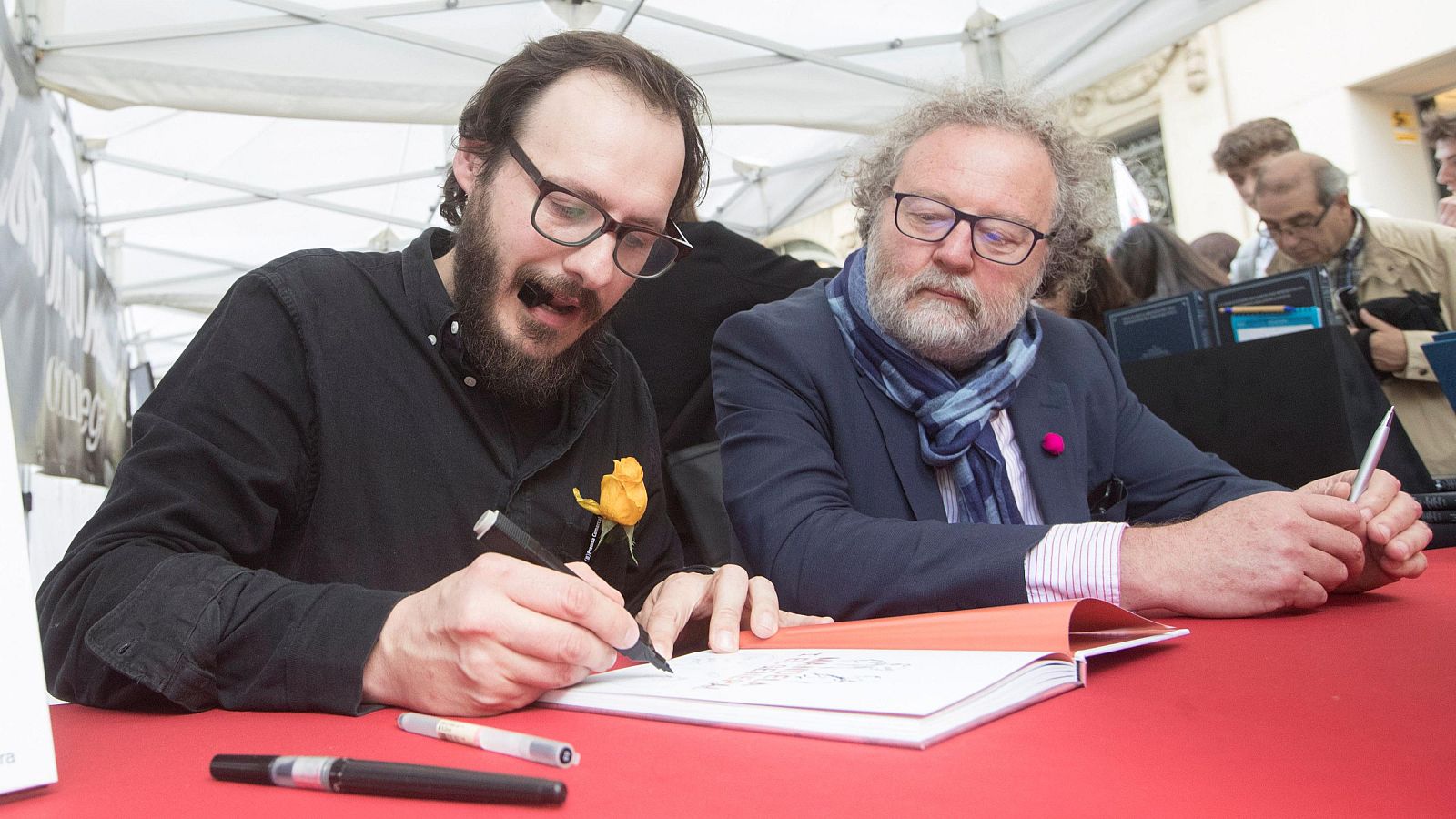 Firma de libros: Dos hombres con gafas firman un libro abierto sobre una mesa con mantel rojo, rodeados de bolígrafos.  Al fondo, se observa un grupo de personas.