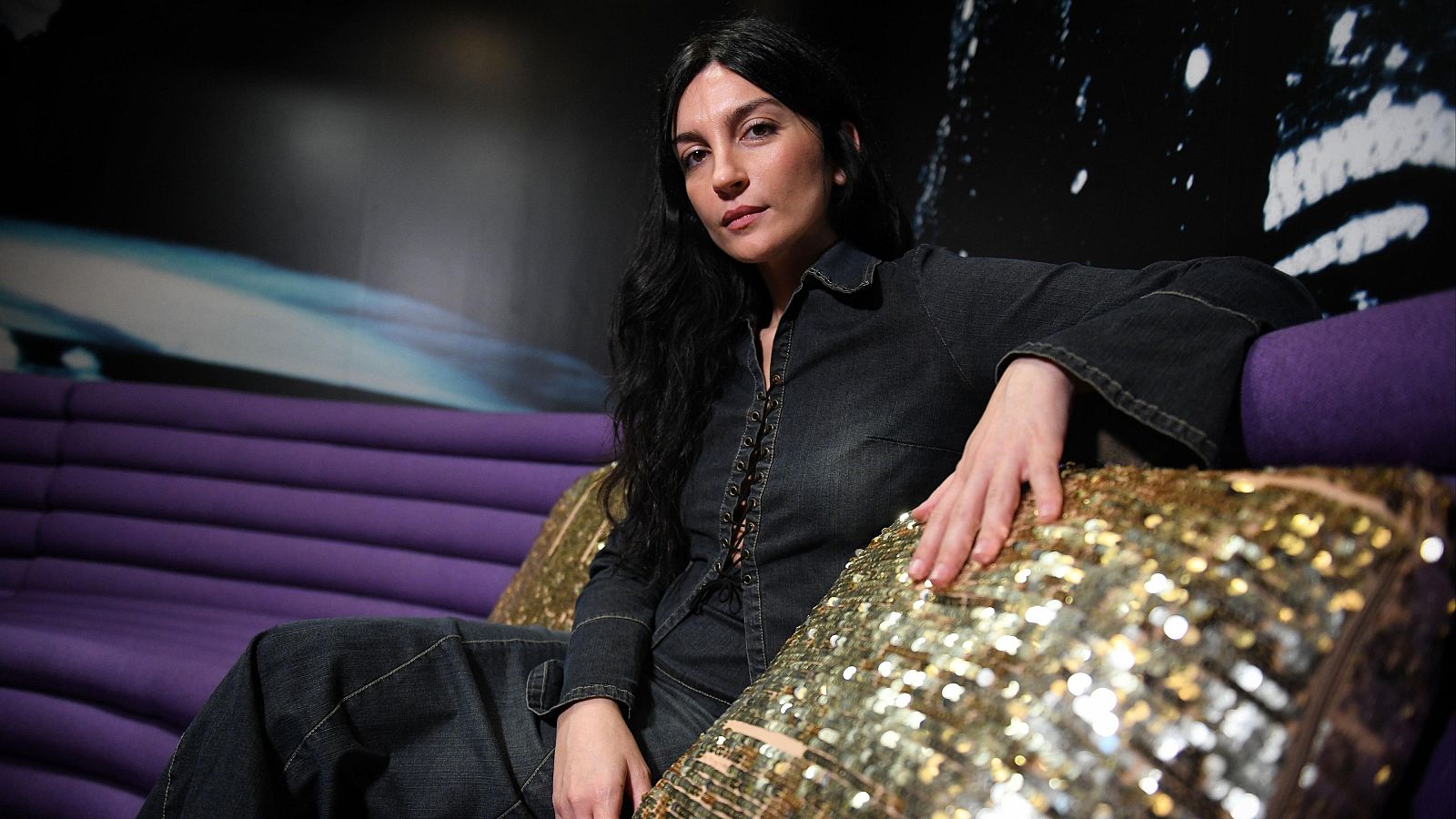 Cantante con camisa vaquera sentada en un sofá morado durante una entrevista.  Expresión seria, mirando a cámara. Fondo con imagen abstracta.