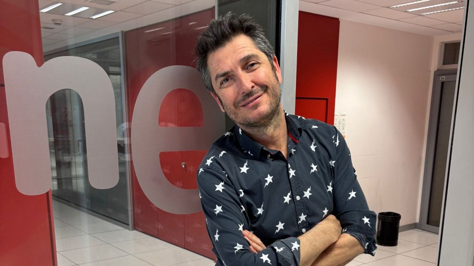 Periodista de RNE con camisa negra de estrellas blancas, brazos cruzados y leve sonrisa, en pasillo o estudio con logo de RNE al fondo.