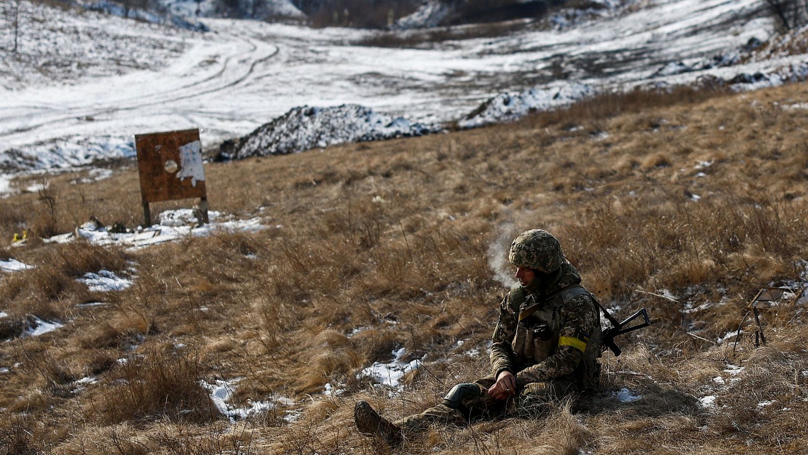 Soldado ucraniano en descanso, fumando, con uniforme de camuflaje y brazalete amarillo.  Rifle cercano, entorno invernal con nieve y hierba seca.