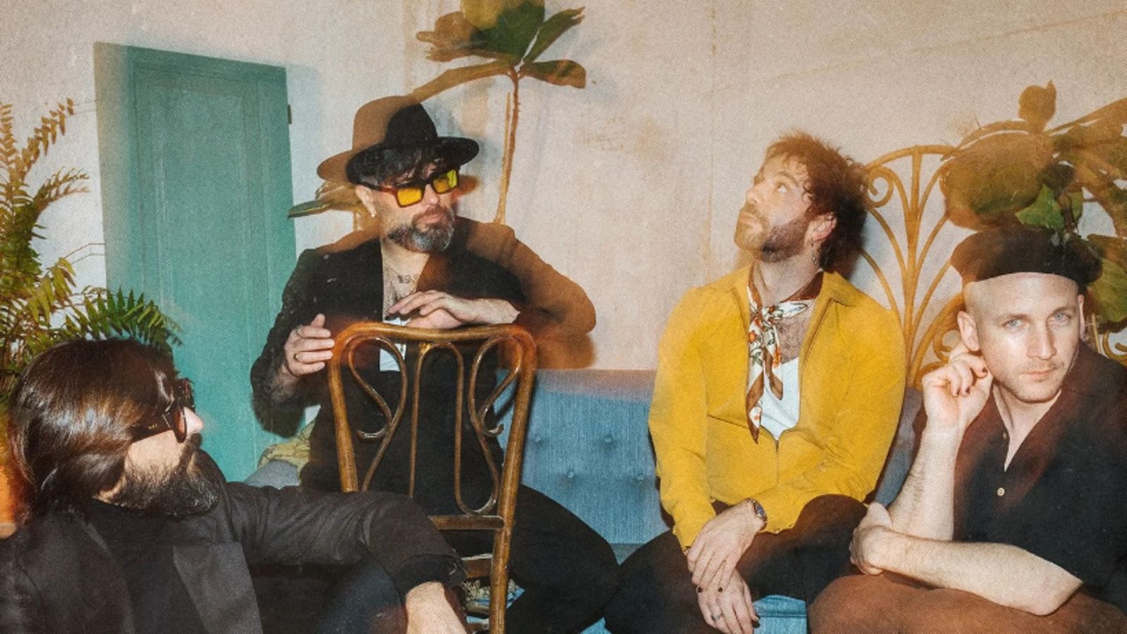 Cuatro hombres con atuendos retro, gafas de sol y sombreros, en un interior con plantas y un sofá. Posible portada de álbum musical.