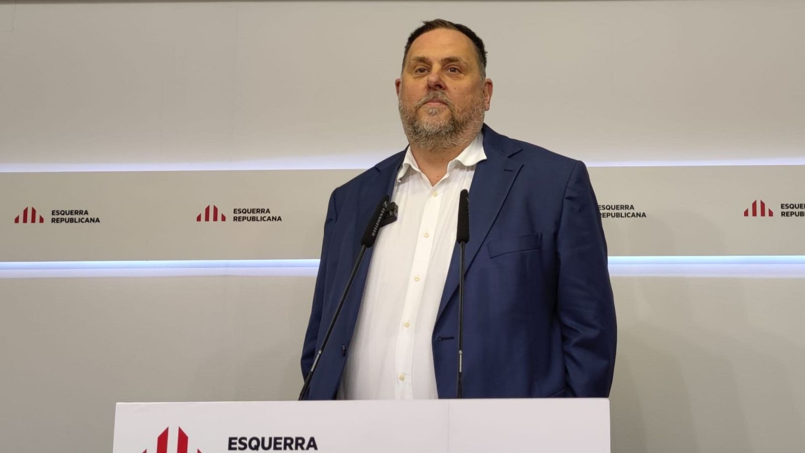 Rueda de prensa de Oriol Junqueras tras un atril con micrófono. Americana azul marino y camisa blanca. Expresión seria.