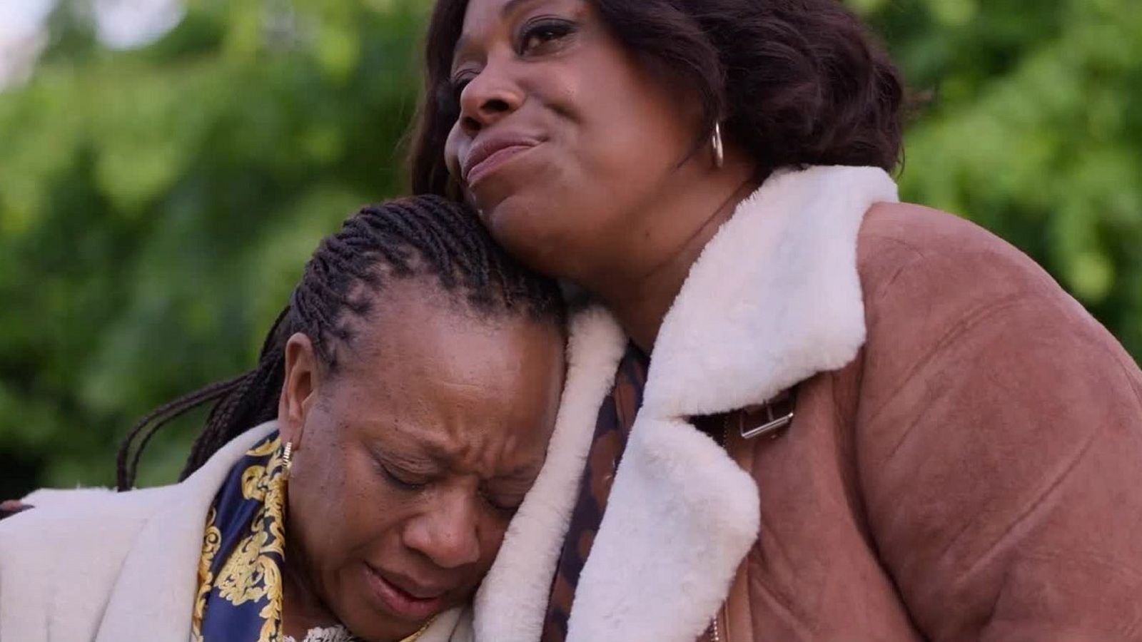 Abrazadas en un parque, dos mujeres de piel oscura expresan tristeza; una llora, la otra la consuela.  Profunda intimidad y dolor compartido.