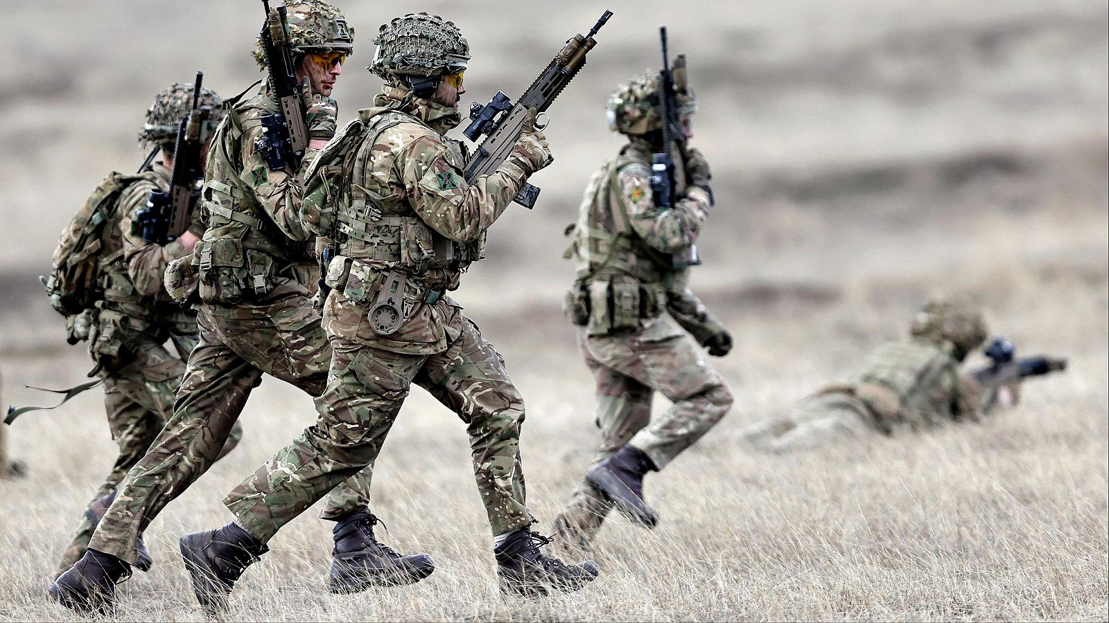 Ejercicio militar: soldados en uniforme de camuflaje avanzan por terreno abierto, algunos con armas listas. Uno yace en el suelo.