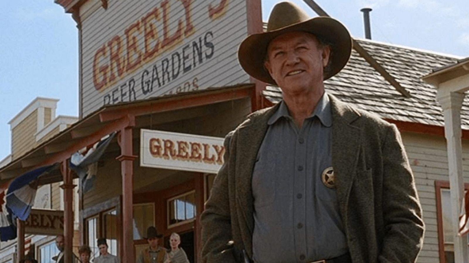 Imagen de Gene Hackman en "Sin Perdón", con atuendo de sheriff (sombrero, chaqueta marrón, camisa gris y estrella) frente a un edificio de madera con letrero "GREELY'S DEER GARDENS".