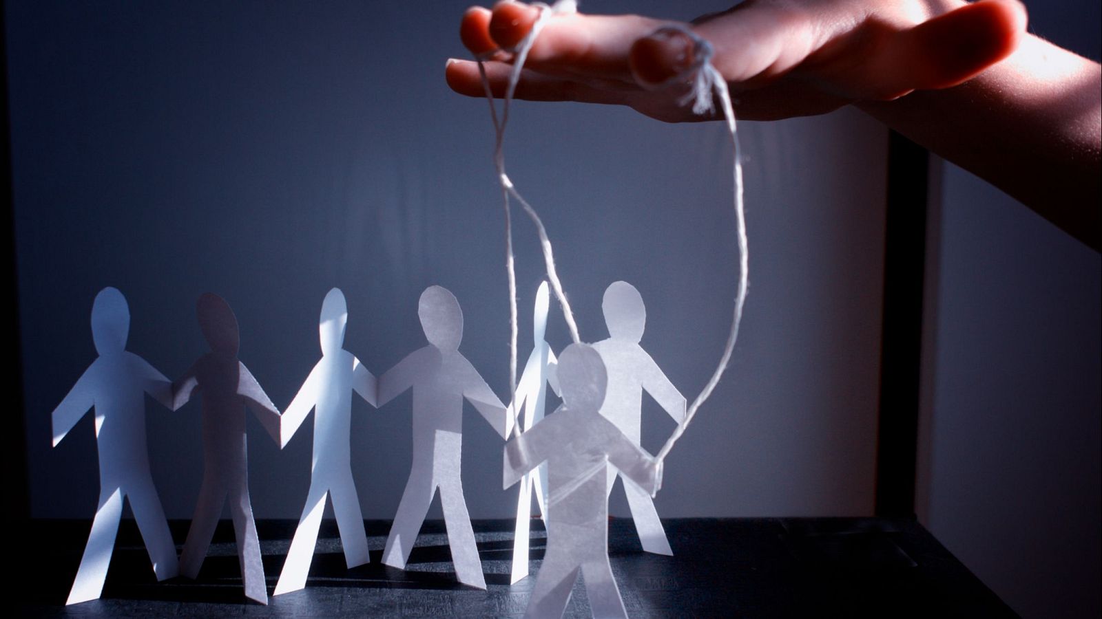 Figuras de papel recortadas, un grupo unido y una sola figura manipulada por hilos desde arriba. Representación visual de control e influencia.