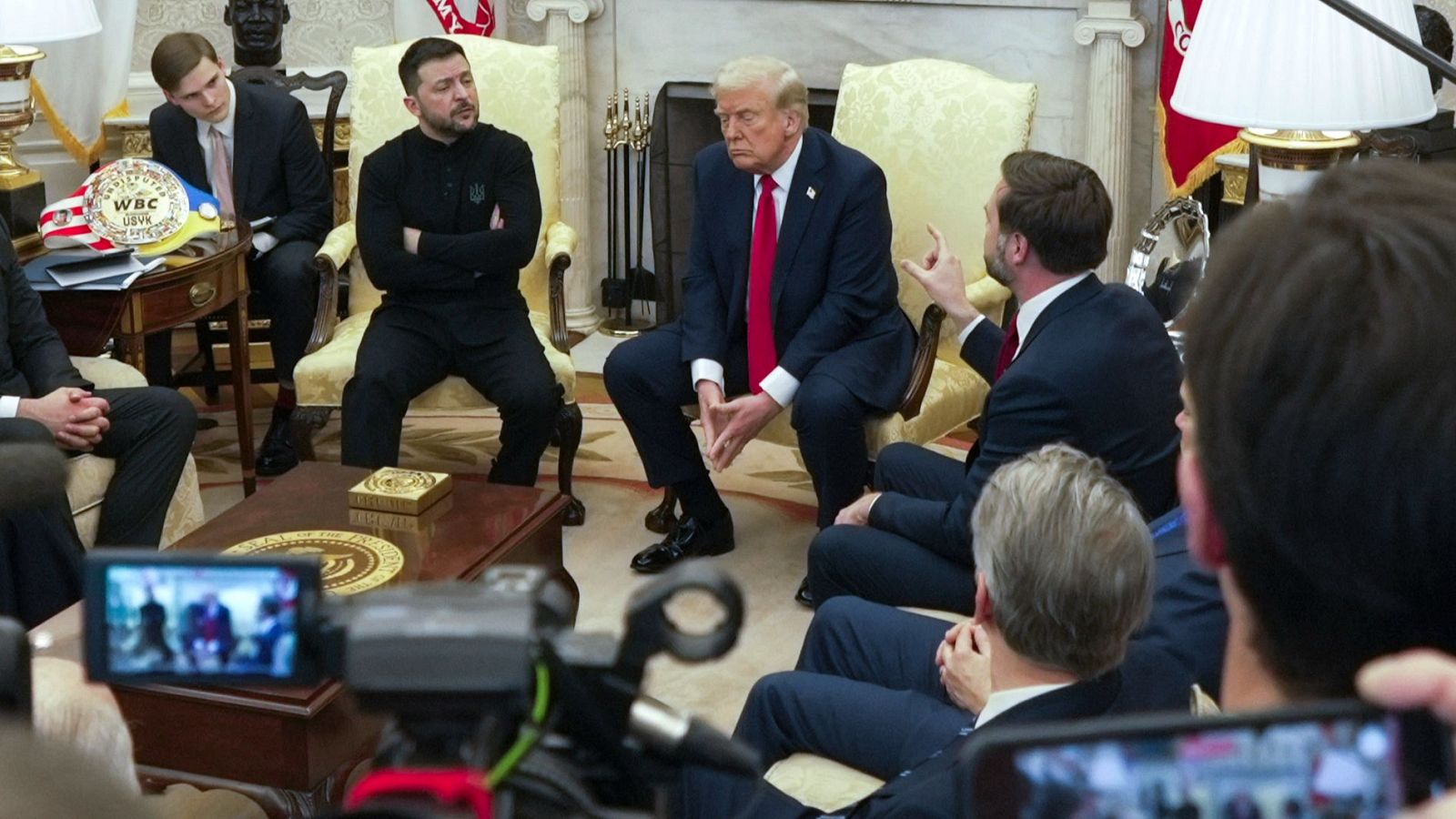 Reunión seria entre dos líderes, uno en traje y otro en ropa informal.  Cámaras presentes. Objeto sobre mesa.