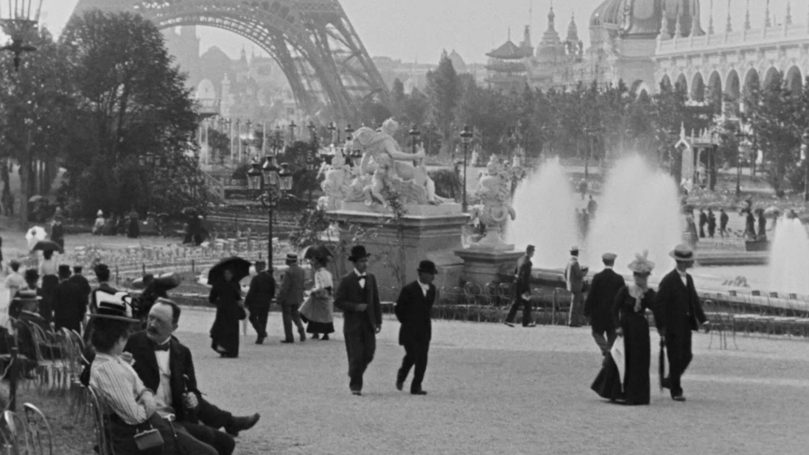 Escena callejera parisina en blanco y negro con multitud de personas elegantemente vestidas, cerca de la Torre Eiffel. Atmósfera festiva y soleada.