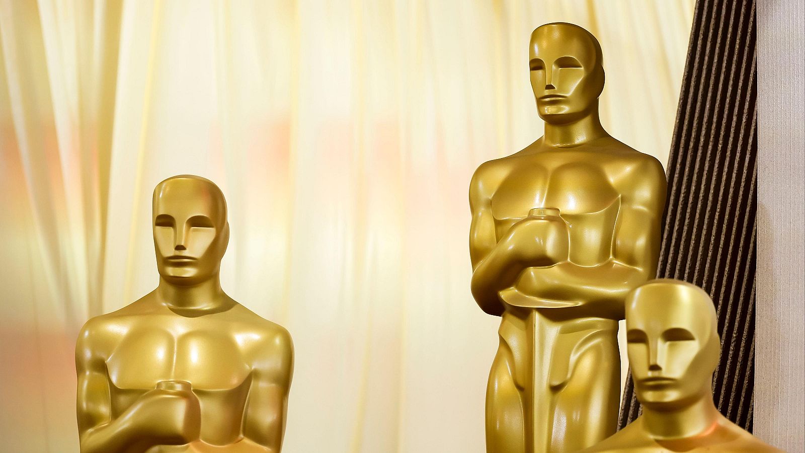 Tres réplicas doradas de los premios Oscar, figuras humanas estilizadas con los brazos cruzados, sobre fondo de tela dorada y marrón oscuro.