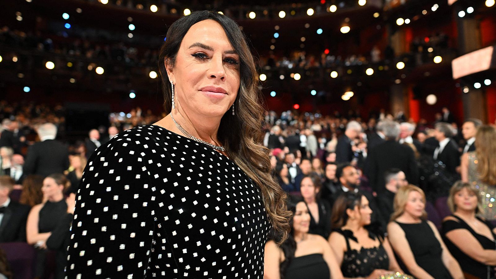 Primer plano de Karla Sofía Gascón en los Premios Oscar; viste un vestido negro con lunares blancos y sonríe ligeramente a cámara. Fondo: auditorio iluminado.