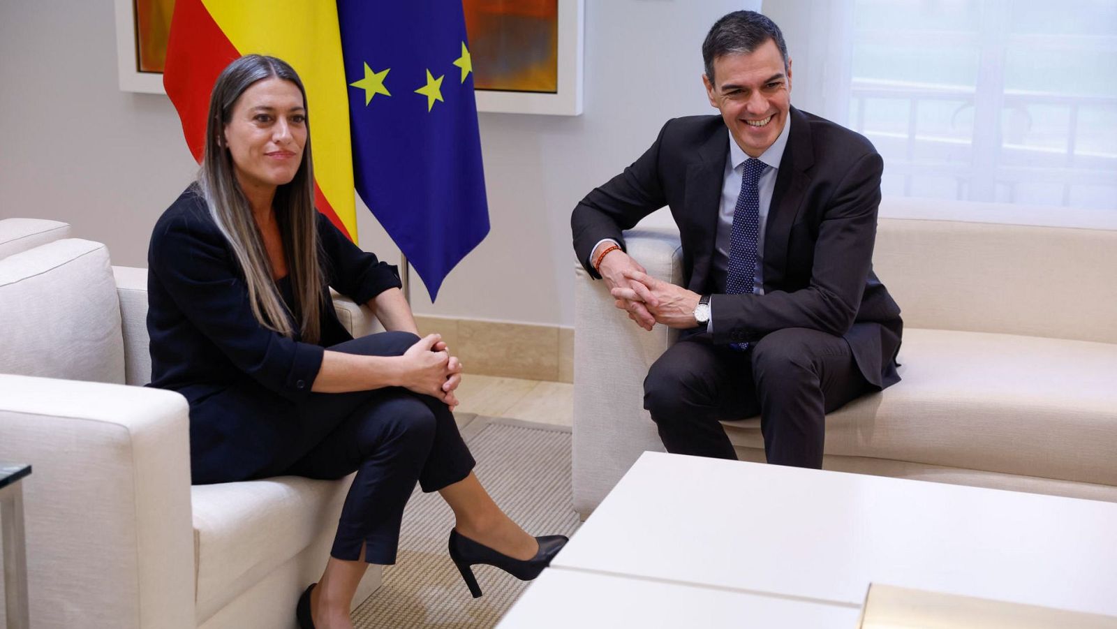 Reunión entre dos personas en sillones beige, con banderas de España y la UE al fondo.  Ambas personas visten ropa formal.