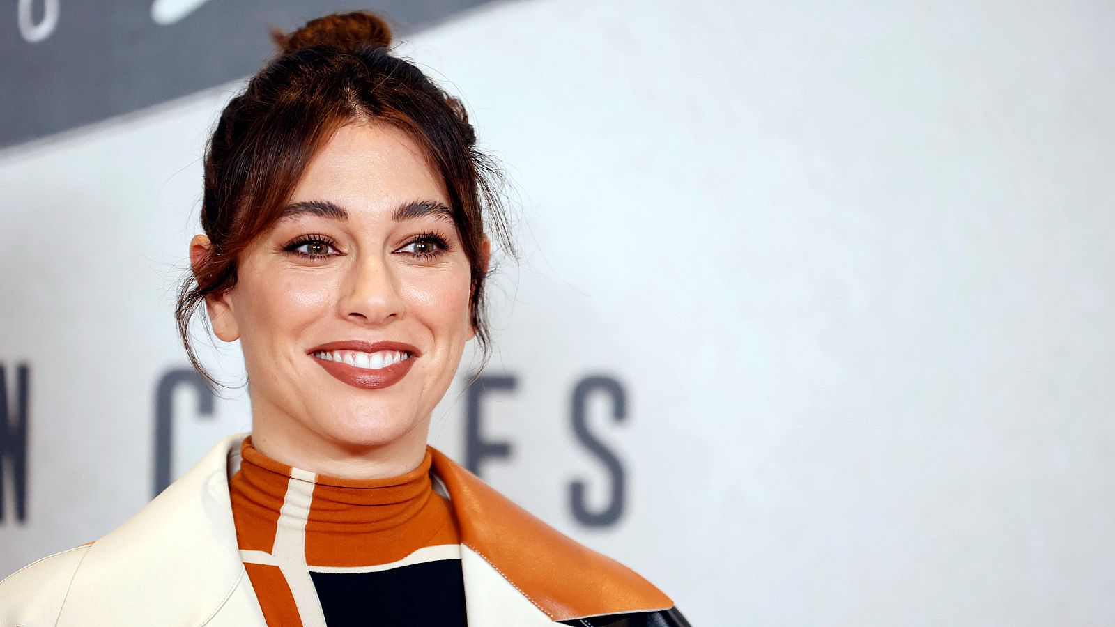Blanca Suárez en la presentación de una película. Luce un moño alto, maquillaje con labios marrón-rojizo y un atuendo con estampado naranja y negro bajo una chaqueta clara.