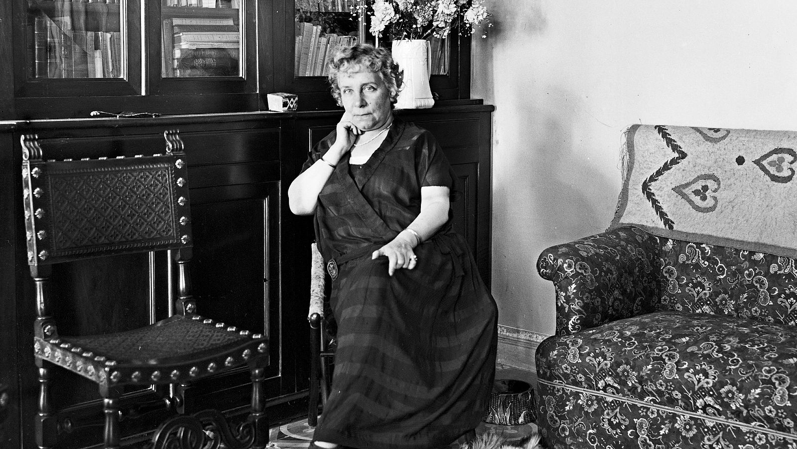 Retrato antiguo en blanco y negro de una mujer sentada, con vestido oscuro y cabello corto.  El ambiente es hogareño, con muebles y flores visibles.