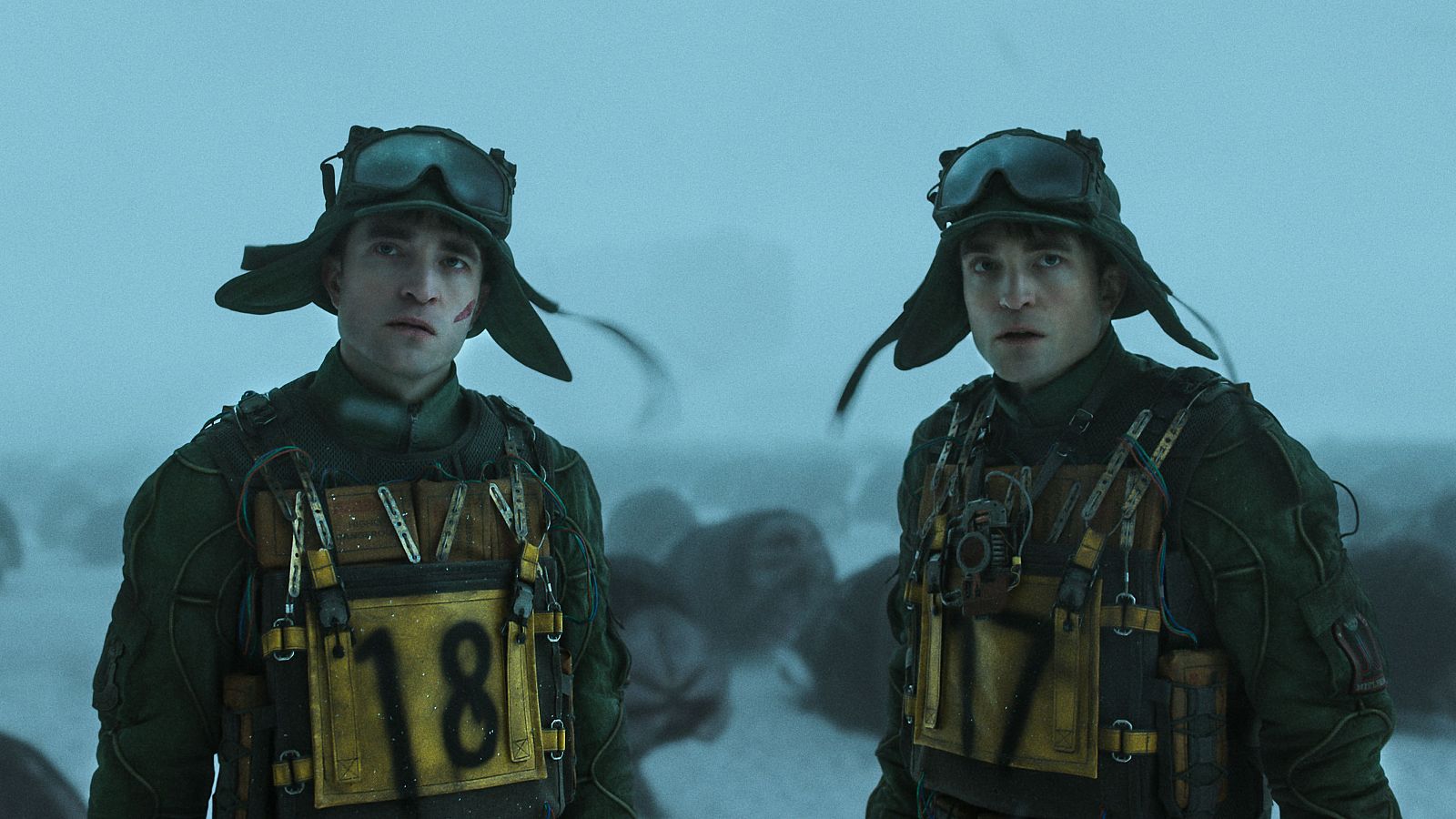 Dos personajes idénticos, con uniformes verdes y placas numeradas, en un paisaje nevado.  Gafas de aviador y sombreros.  Fotograma de película.