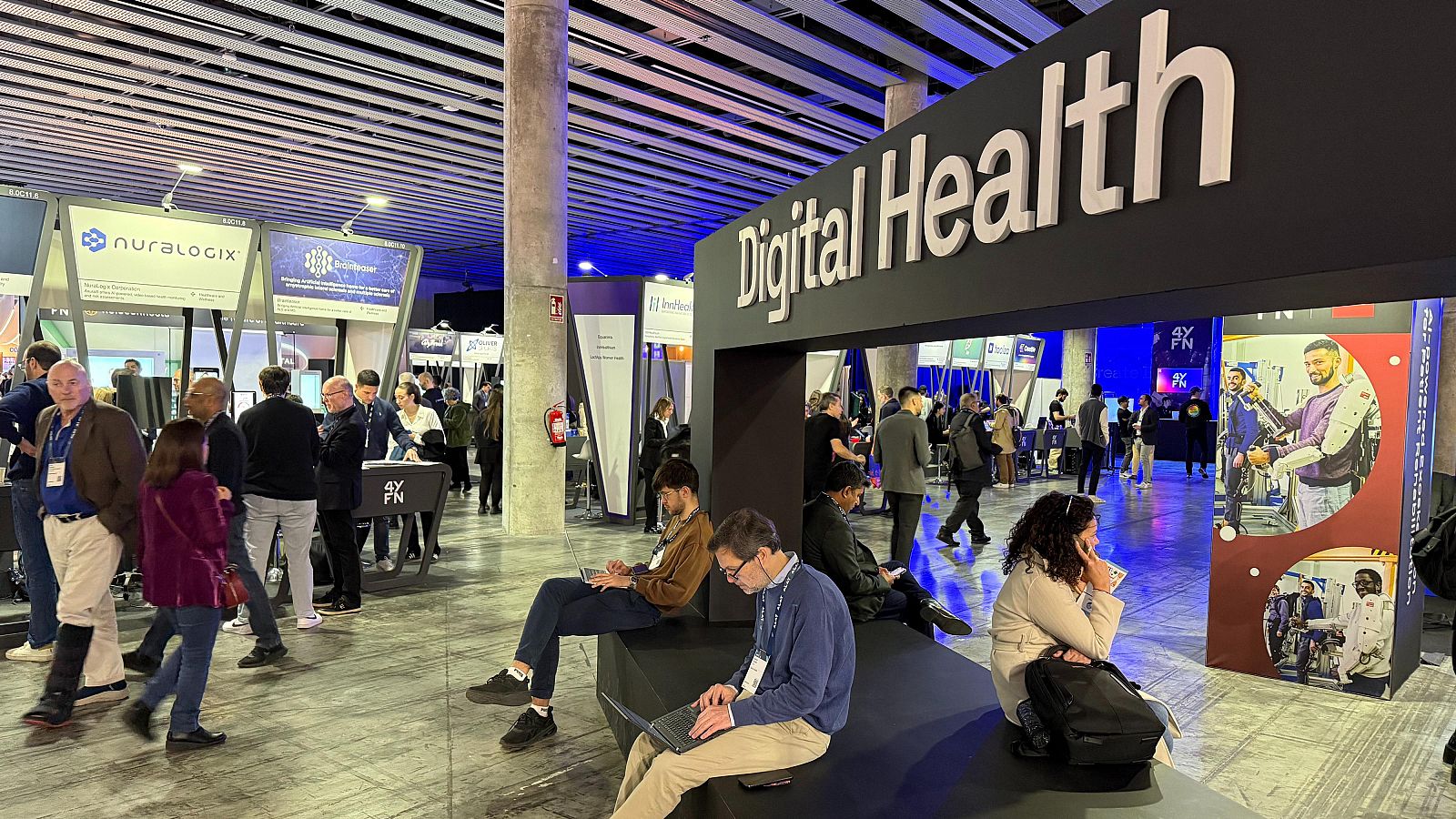 El área de salud digital del MWC muestra asistentes, stands con logotipos empresariales y carteles con imágenes de tecnología médica, como un exoesqueleto robótico, reflejando la innovación en este sector.