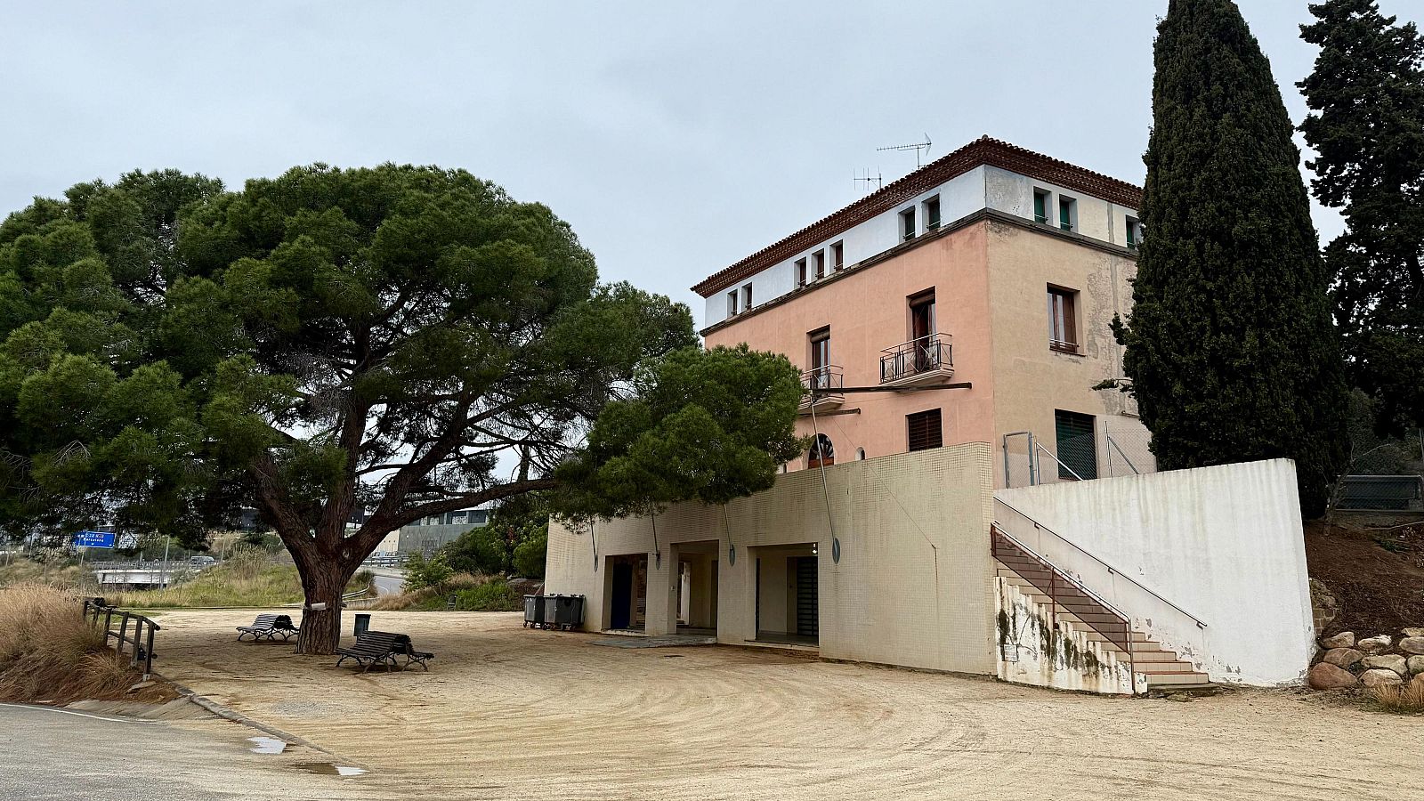 Vista lateral de un edificio rosa salmón con detalles blancos, escaleras exteriores y una extensión beige en la planta baja. Árboles y terreno arenoso rodean la estructura.