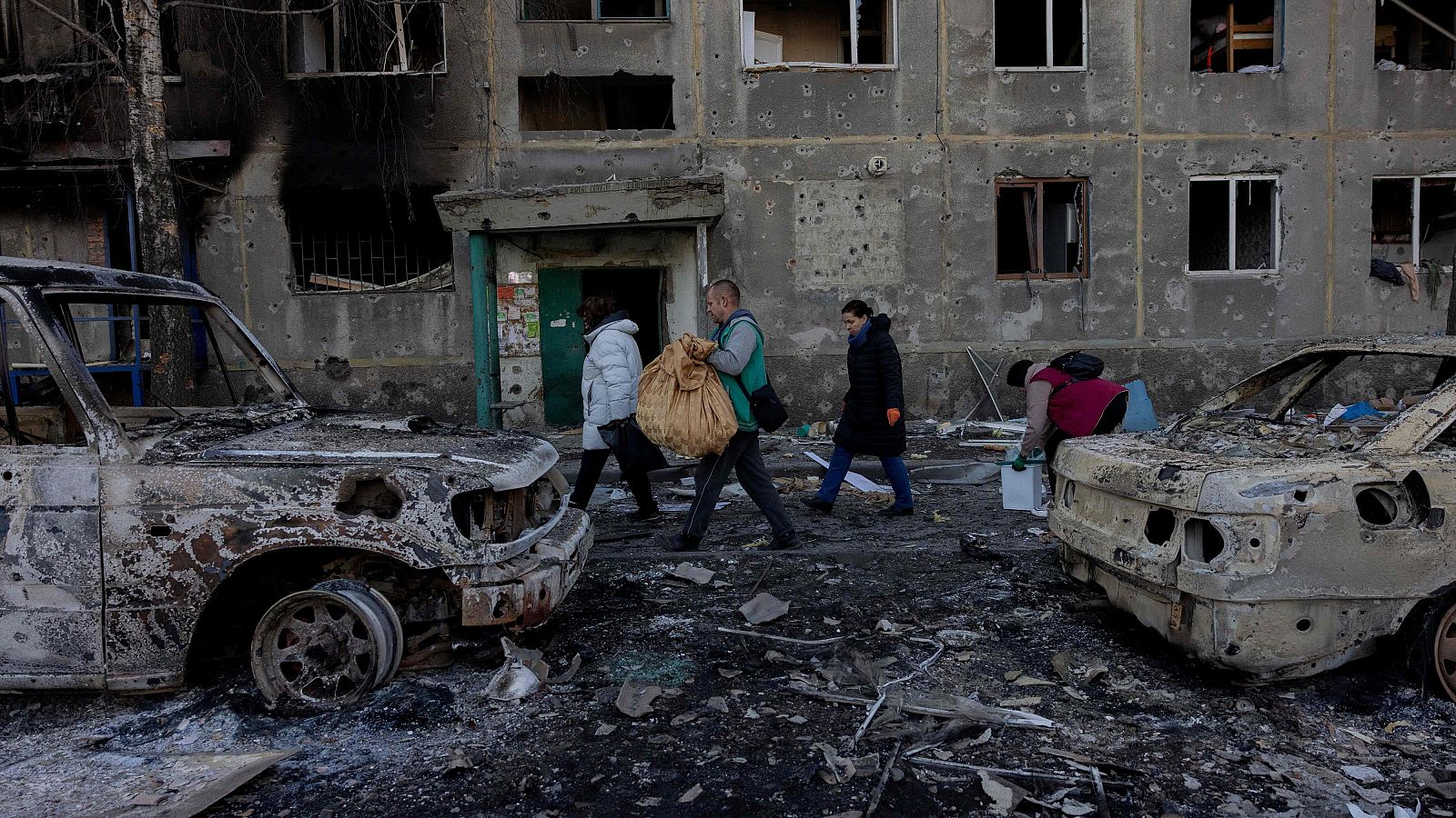 Calle ucraniana devastada por la guerra: edificio dañado, coches calcinados, cuatro personas entre escombros, una recogiendo objetos.