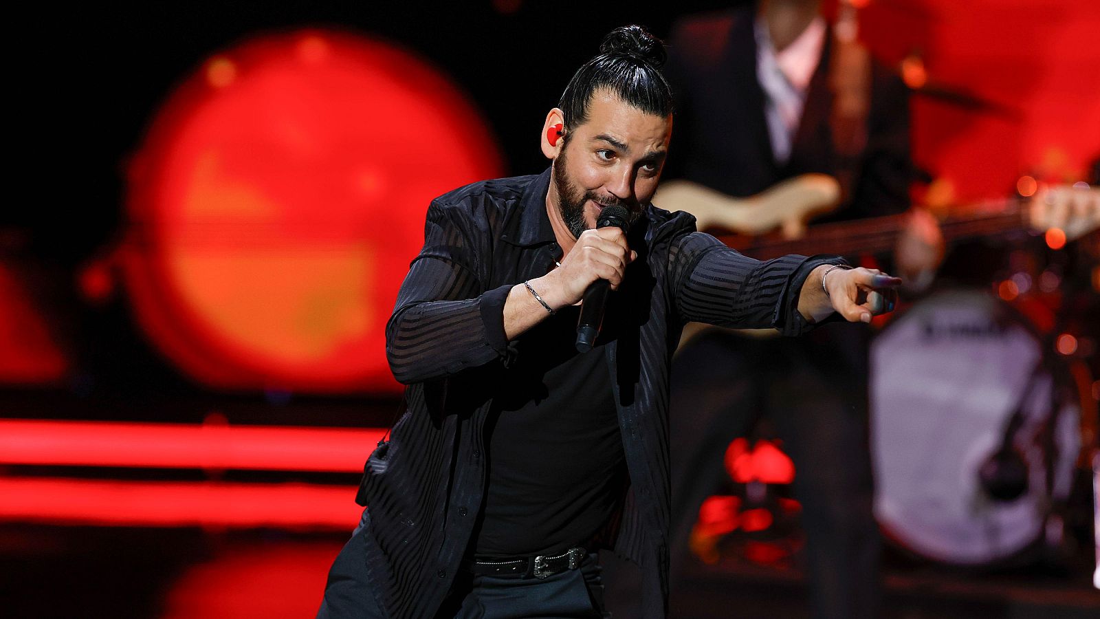 Artista masculino en escenario con micrófono, camisa negra a rayas y pantalón oscuro, actuando en una gala con iluminación roja y banda musical al fondo.