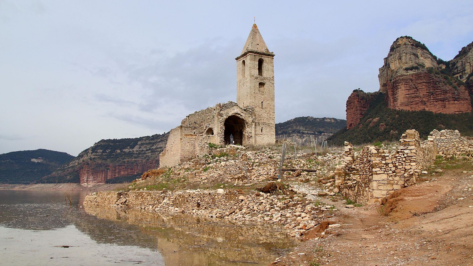 Ruinas de una iglesia de piedra clara con campanario, situada en zona rocosa junto a un embalse.  Restos de muros y vegetación escasa.