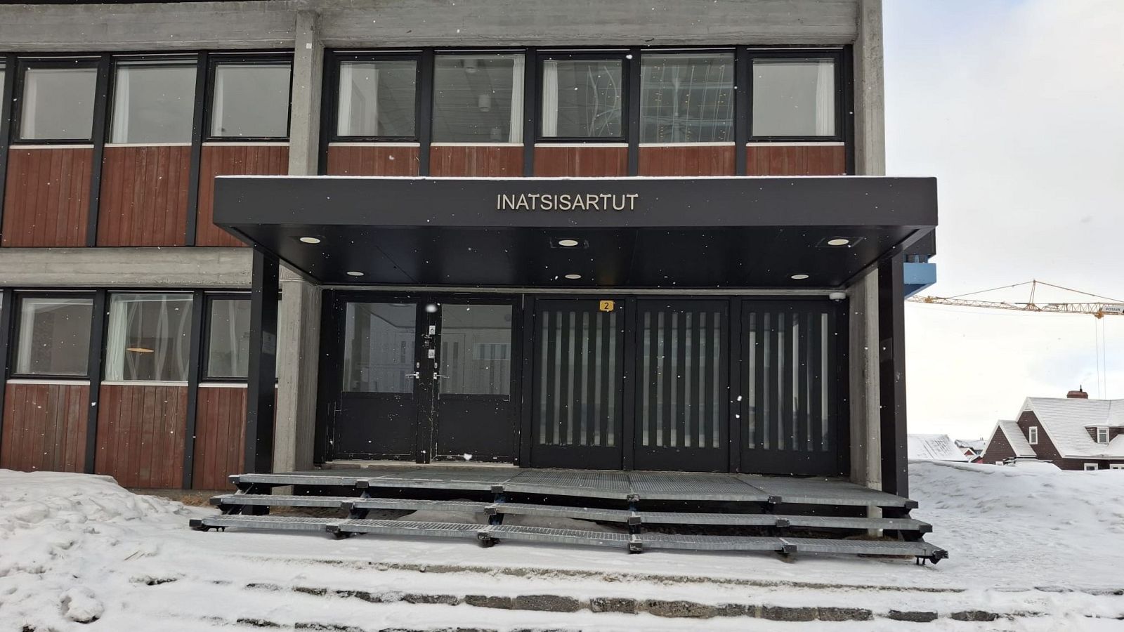 Entrada al Parlamento de Groenlandia en un día nevado. Edificio de dos plantas con fachada de cemento y madera, acceso con escalones metálicos y un tejado protector.