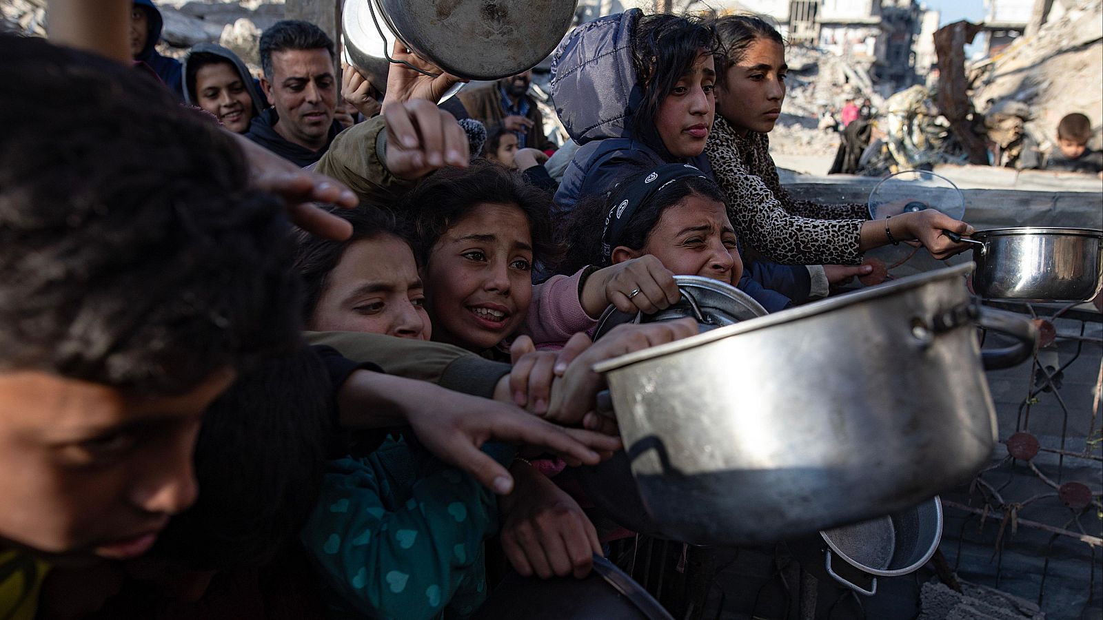 Personas, mayormente niños, esperan comida con expresiones de necesidad y angustia en un entorno destruido.