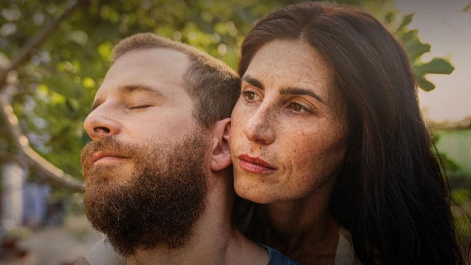 Pareja abrazándose al aire libre; él, con barba pelirroja y ojos cerrados; ella, con cabello oscuro, pecas y expresión seria mirando a la derecha. Intimidad y melancolía.