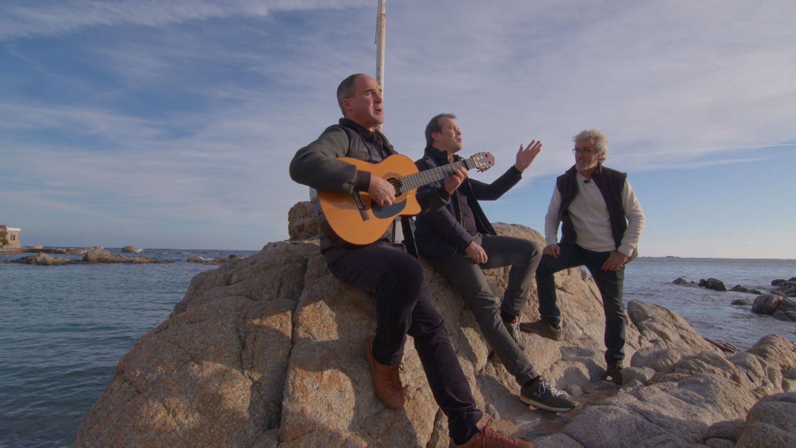 Actuación musical en costa rocosa con tres hombres: un guitarrista, un cantante y un posible presentador.  Se observa el mar al fondo.