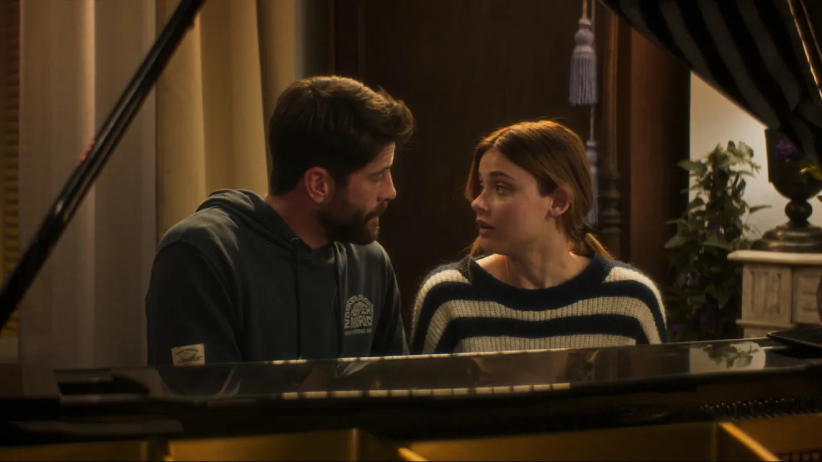 Dos actores, uno con sudadera oscura y otro con jersey a rayas, sentados frente a un piano, en una escena con atmósfera seria.