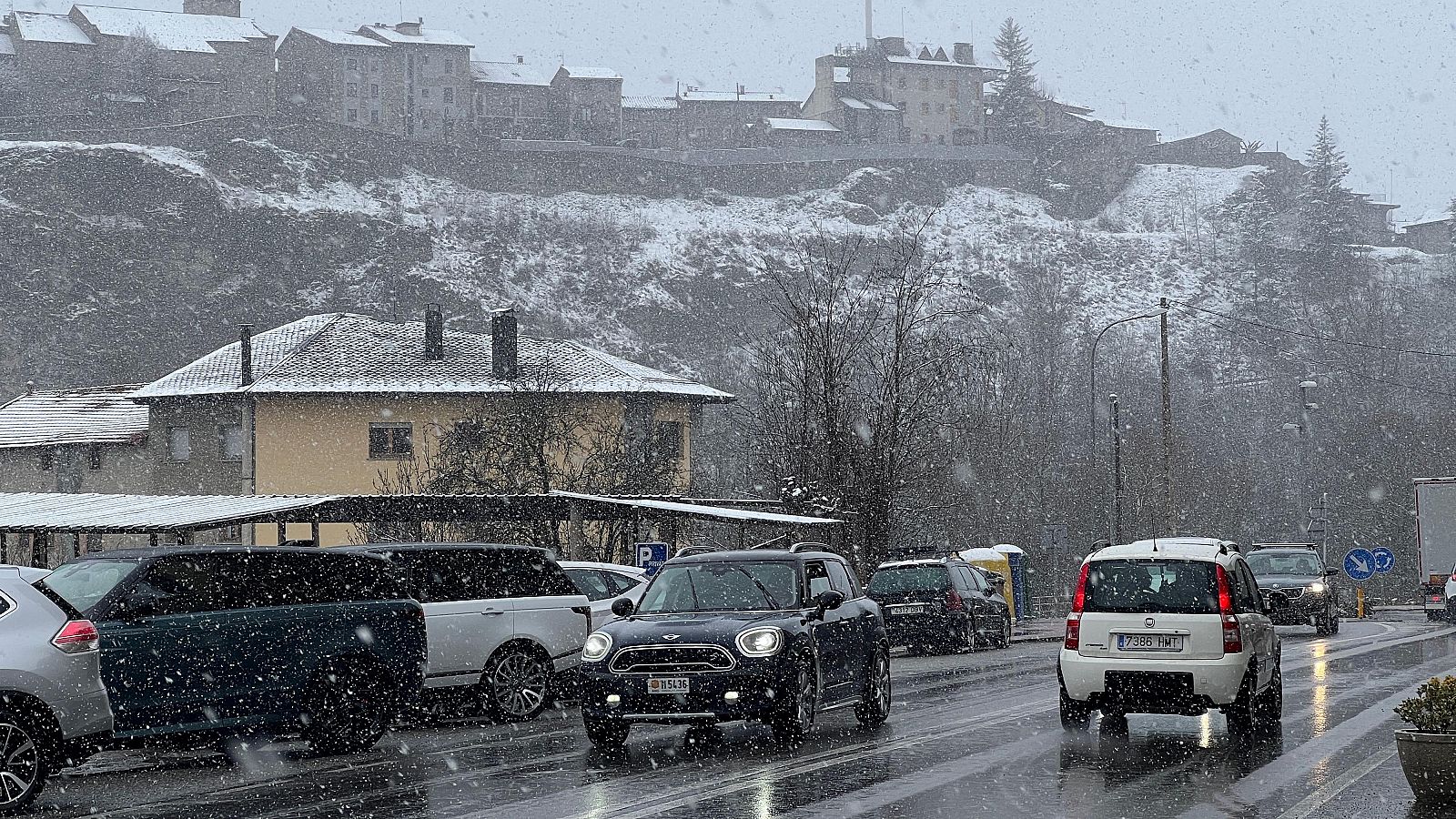 Calle nevada con coches, edificios con tejados cubiertos de nieve y una colina al fondo con casas y una estructura elevada.