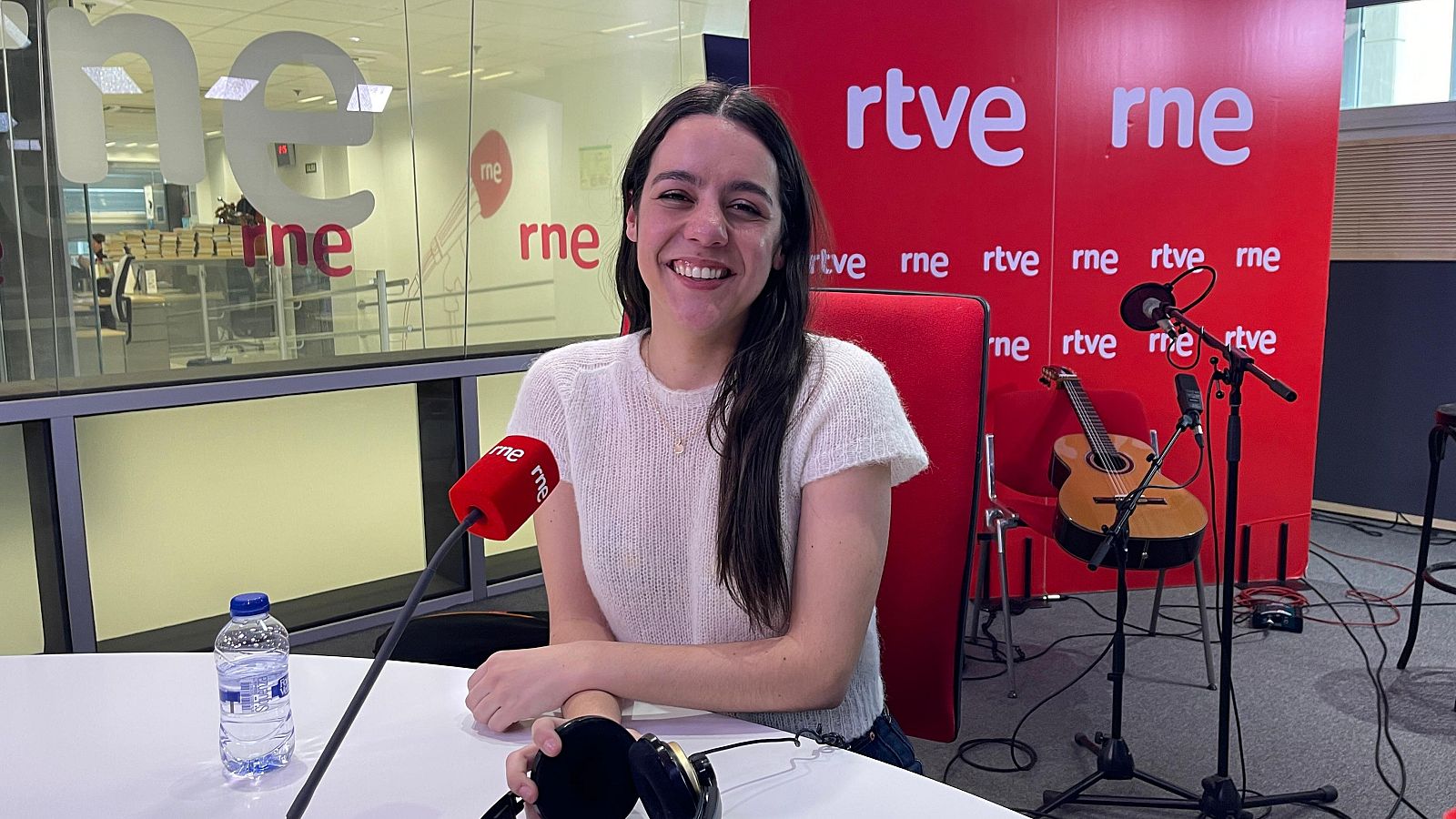 Cantante en estudio de radio RNE, sonriendo ante micrófono.  Guitarra acústica visible. Presenta su nuevo disco.