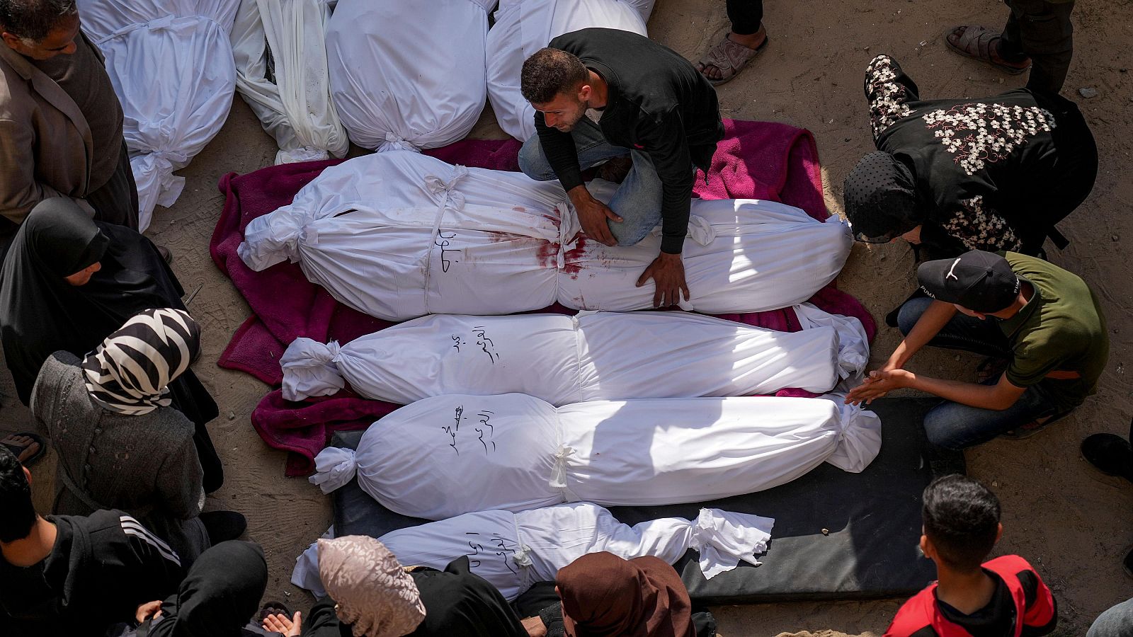 Escena de duelo en Gaza: cuerpos envueltos en sudarios blancos con manchas de sangre sobre tela granate, rodeados de dolientes con rostros de tristeza.