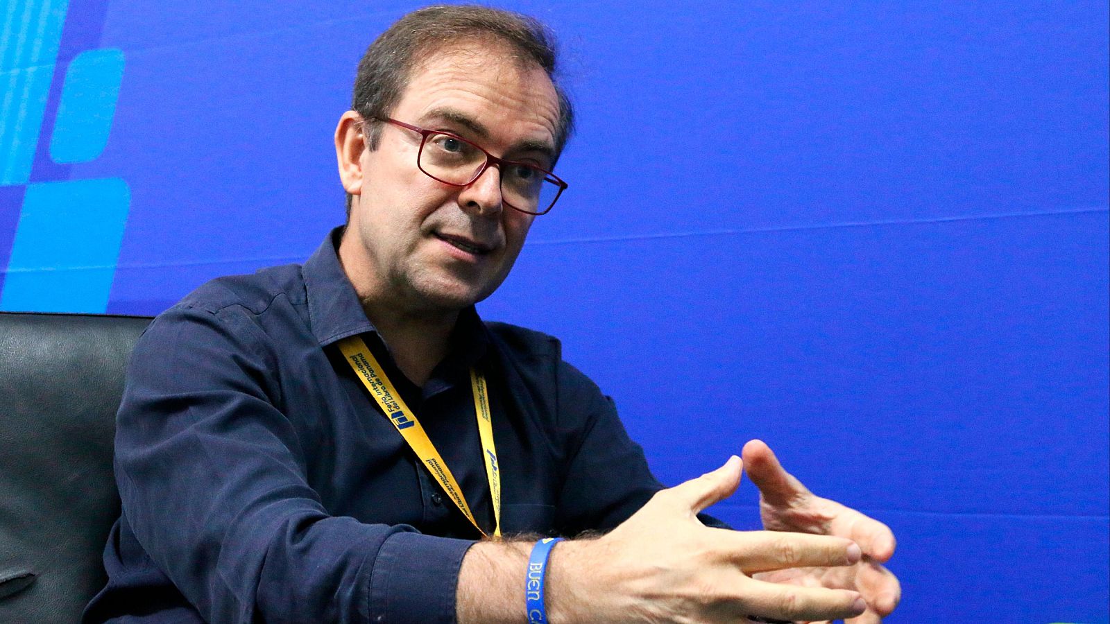 Javier Sierra, en entrevista, gesticula mientras habla. Viste camisa azul oscuro, gafas rojas y una acreditación amarilla.