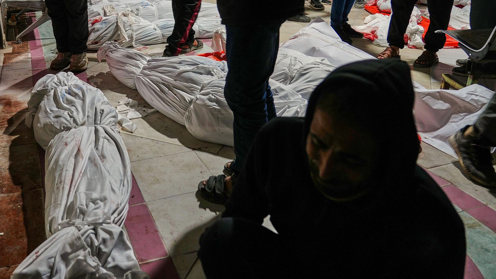 Cuerpos cubiertos con telas blancas yacen en el suelo de un hospital tras un ataque aéreo. Un hombre agachado, con la cabeza cubierta, muestra dolor cerca de los cuerpos.