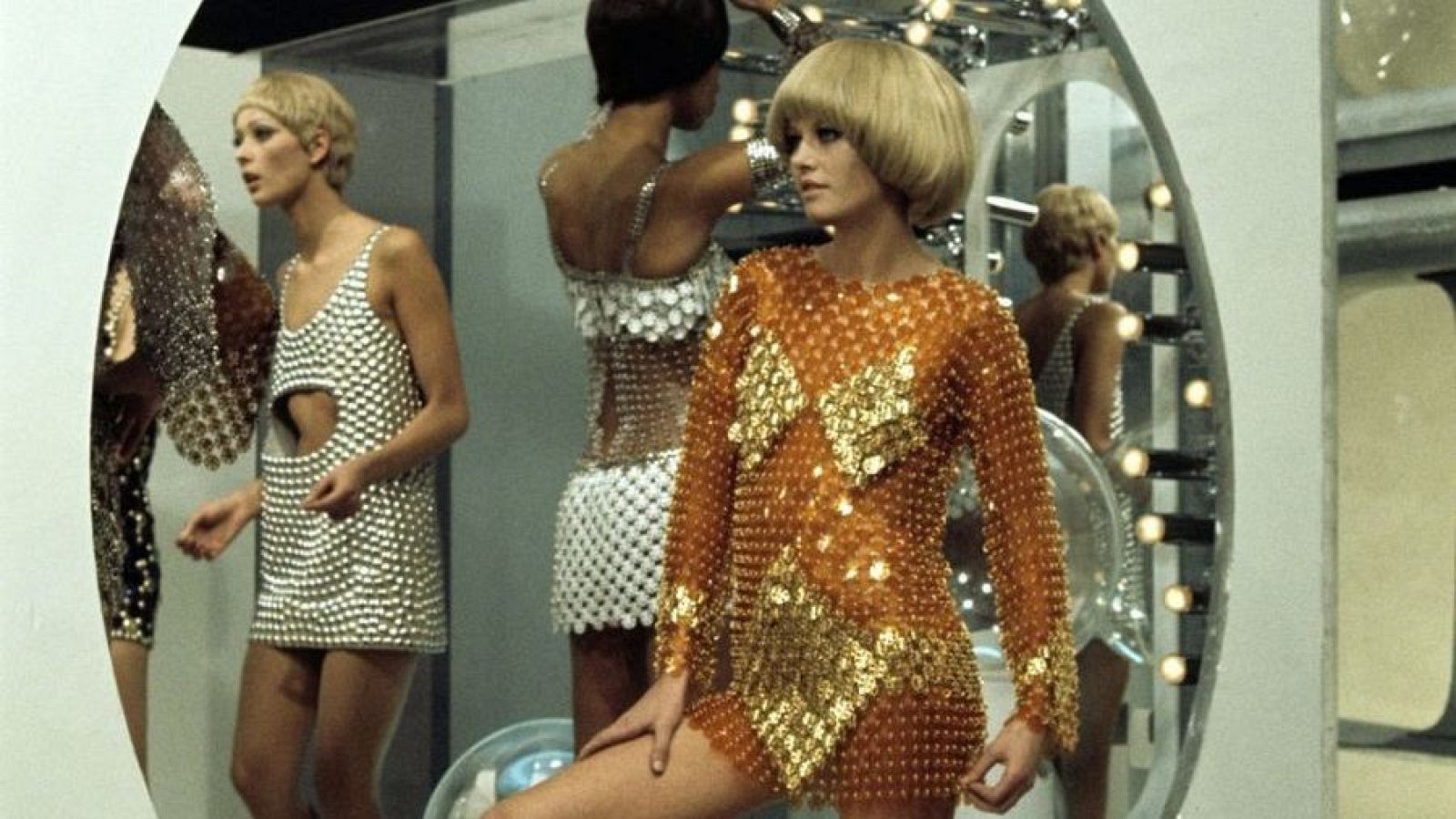 Tres modelos con minivestidos metálicos posan en un camerino; una con vestido dorado, otra plateado y una tercera con un conjunto blanco plateado.