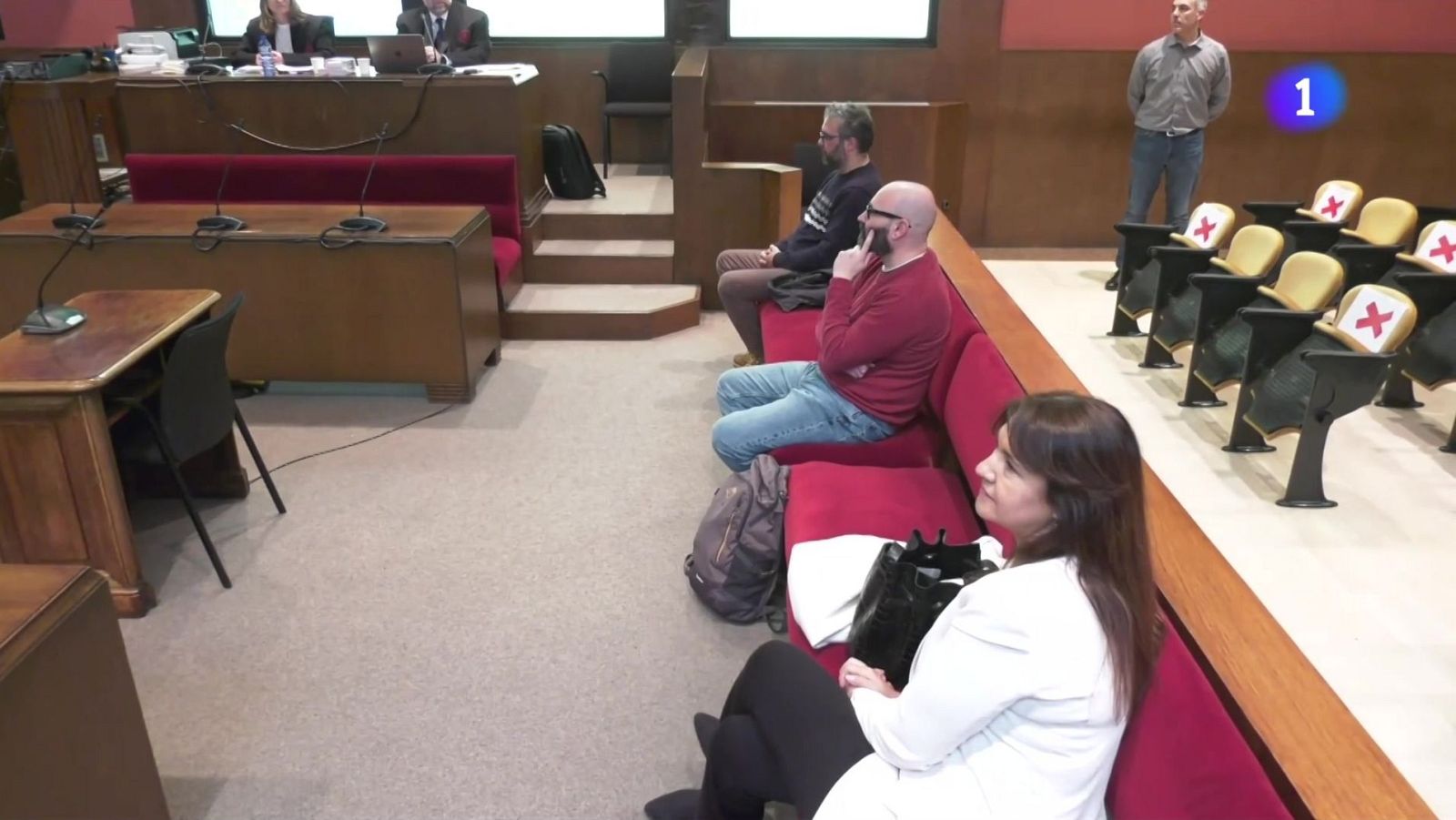 Sala de tribunal con jueces, público en bancos rojos y sillas vacías marcadas con cruces rojas.  Número 1 visible.