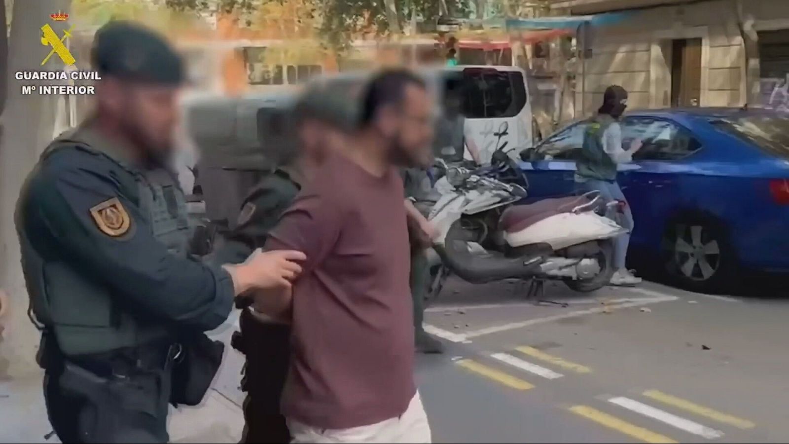 Detención en la calle de un hombre con camiseta granate y pantalones blancos por agentes de la Guardia Civil, con otros agentes presentes.