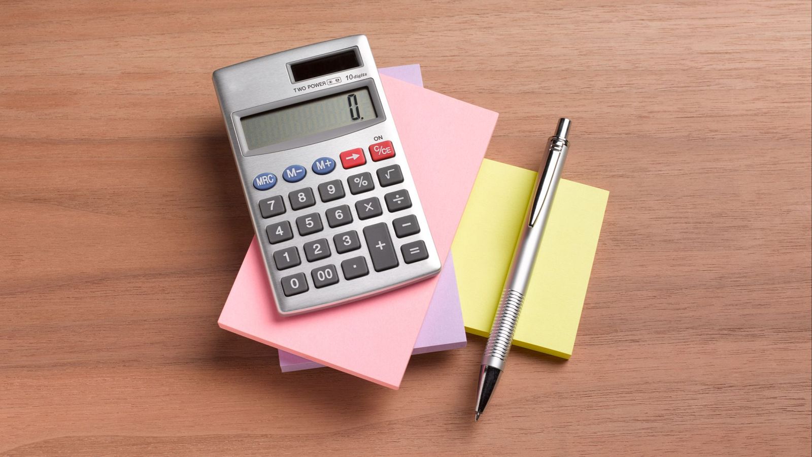 Calculadora plateada encendida mostrando "0", sobre notas adhesivas y bolígrafo plateado, sobre superficie de madera.  Sugiere cálculos.
