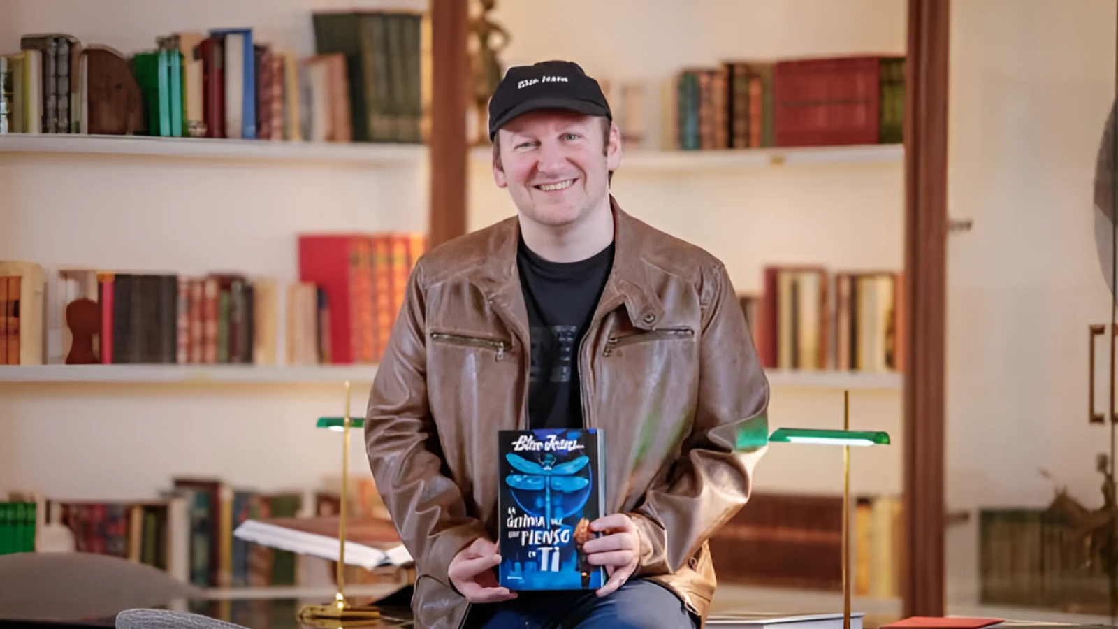 Blue Jeans presenta su nueva novela, "La última vez que pienso en ti", con una portada azul oscura y una libélula. Sonríe mientras sostiene el libro.