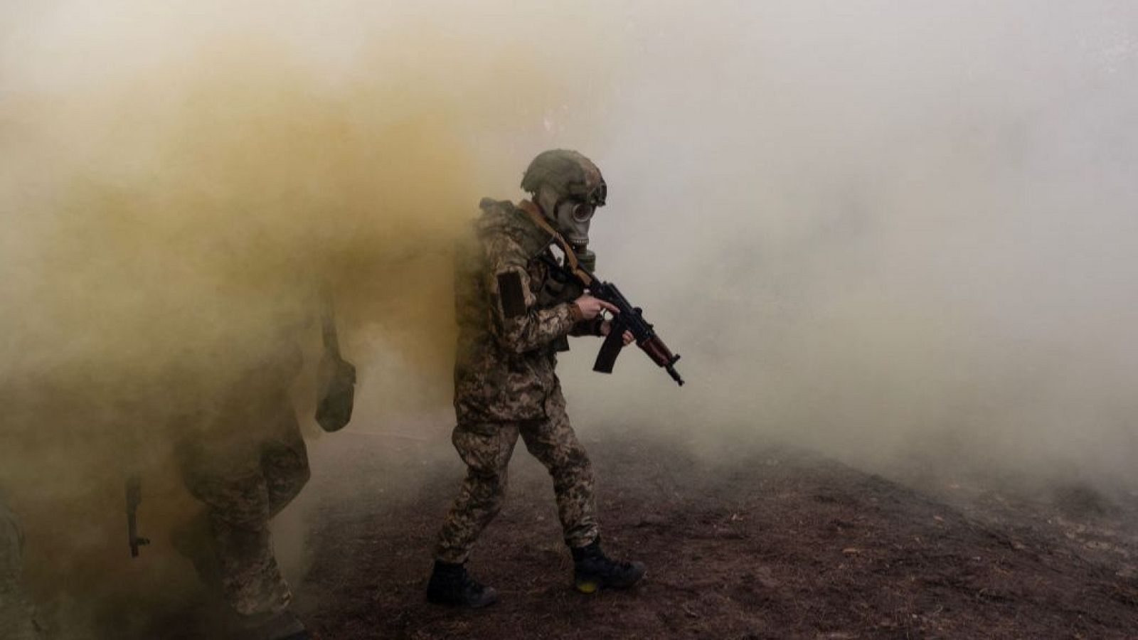 Soldados ucranianos con uniforme de camuflaje y máscaras de gas avanzan entre humo denso, portando rifles de asalto.  Situación de combate o entrenamiento.