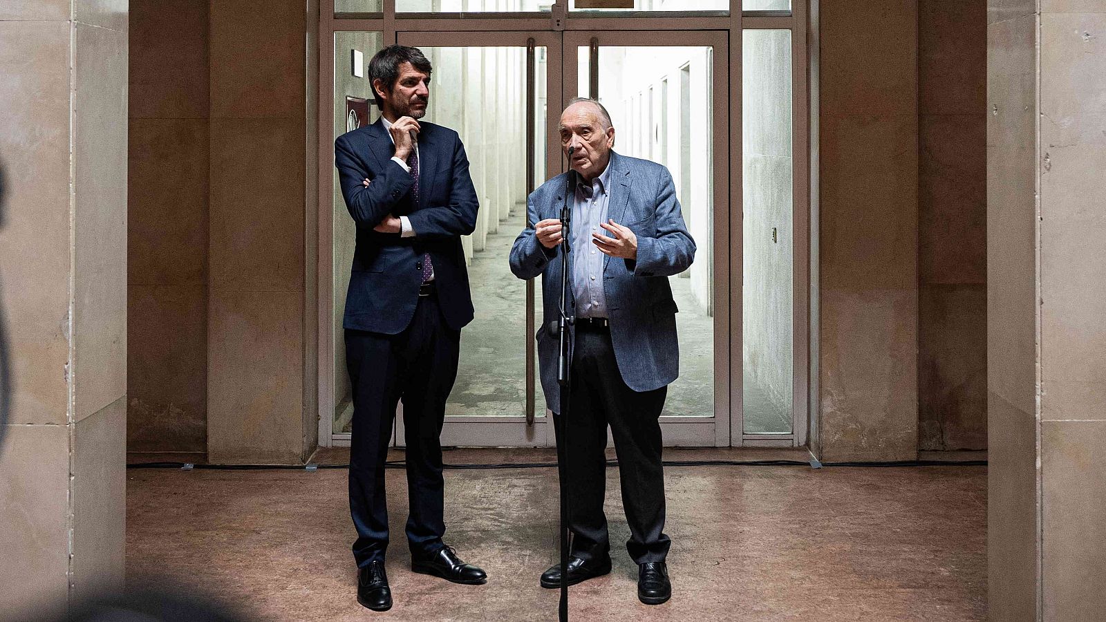 El ministro de Cultura y el presidente de la Academia de Cine conversan; uno habla por micrófono, el otro escucha con los brazos cruzados. Ambos visten formalmente.