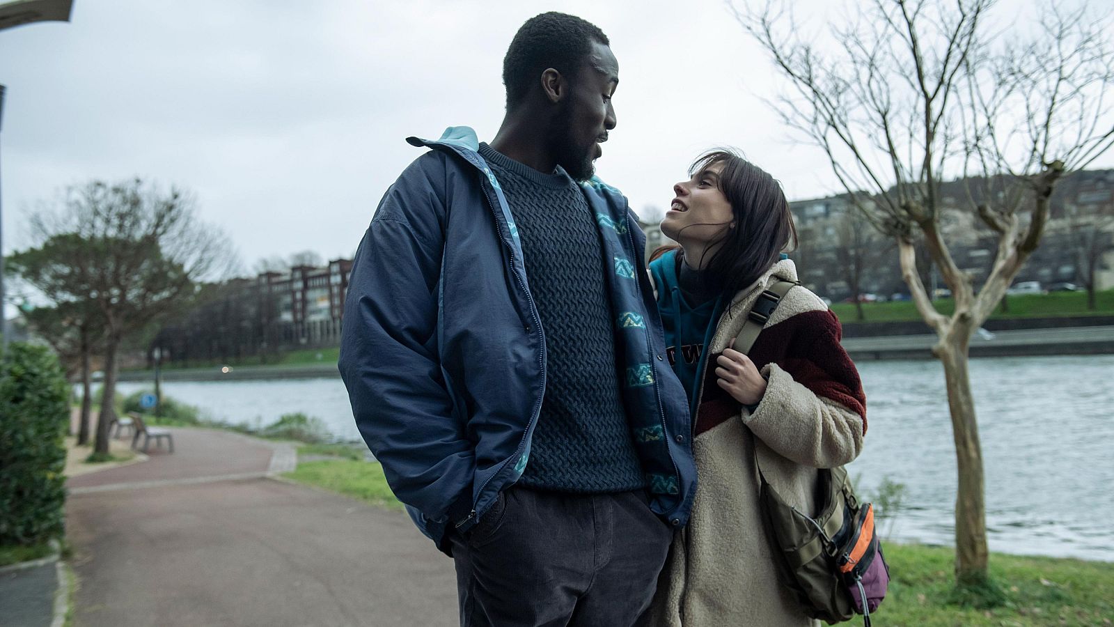 Un hombre y una mujer conversan junto a un río en un día nublado. Él lleva chaqueta azul y jersey verde; ella, abrigo beige y mochila verde oliva.  Ambiente invernal.