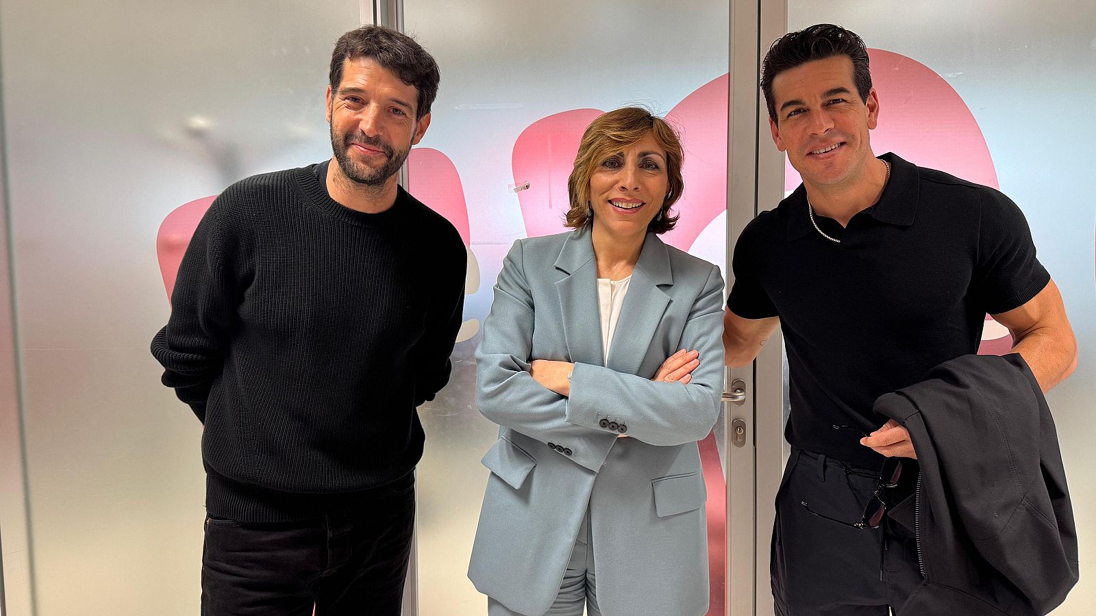 Tres personas, dos hombres y una mujer, presentan una obra. Un hombre viste jersey negro, otro polo negro y la mujer un traje azul claro.