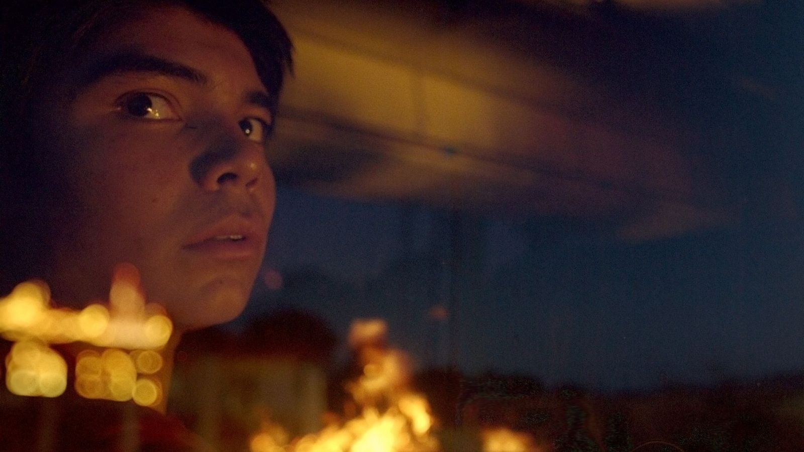 Joven con expresión de sorpresa mirando a través de una ventana o vehículo; luces desenfocadas al frente y fondo borroso con posible fuego o explosión.