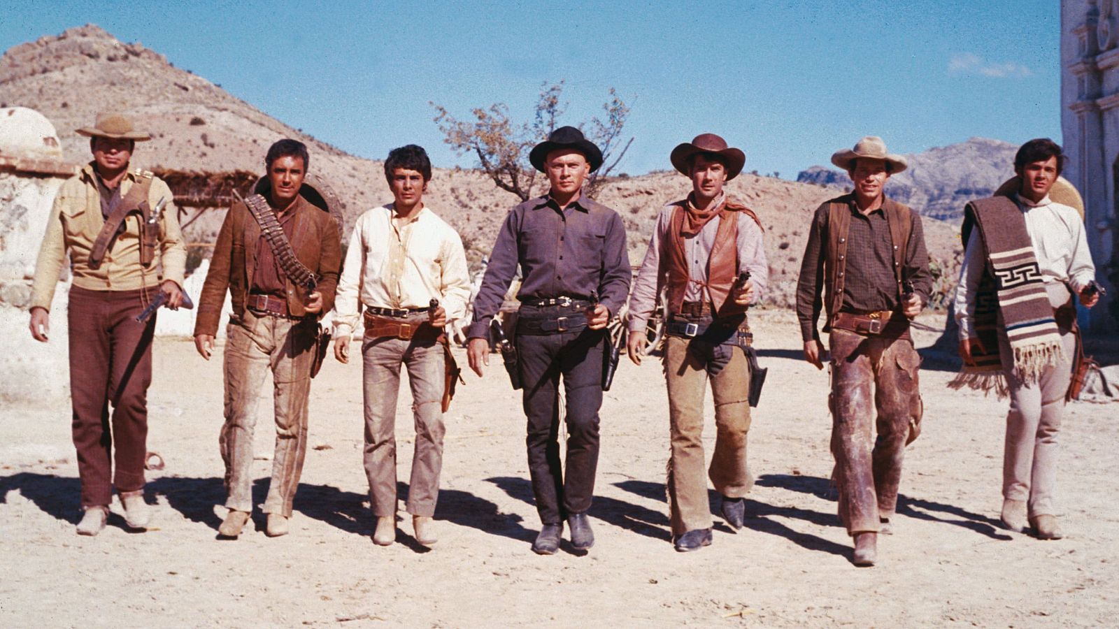 Siete hombres vestidos con atuendos del oeste caminan en fila.  Imagen de una película ambientada en un paisaje desértico.