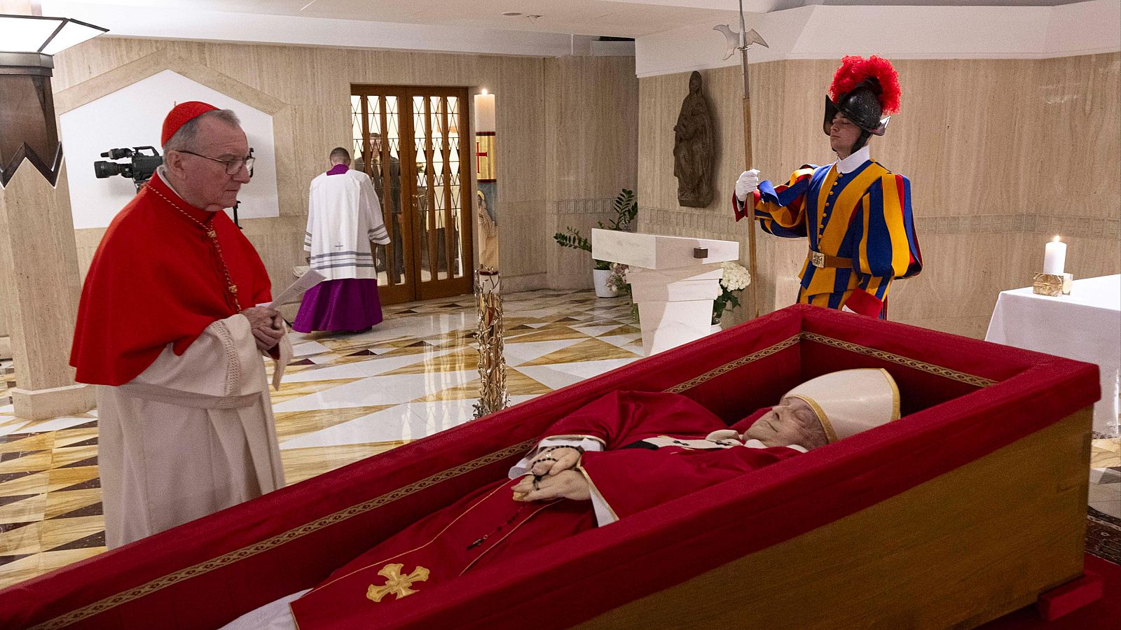 Constatación de la muerte del Papa en la Capilla de Santa Marta.  Un cardenal y un guardia suizo rodean el féretro rojo.  Se observa una cámara de televisión.