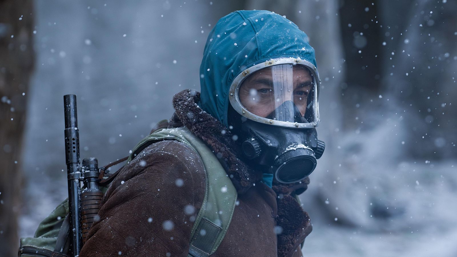 Actor con capucha, máscara antigas y mochila (con arma visible) en escena nevada de bosque.  Chaqueta marrón.