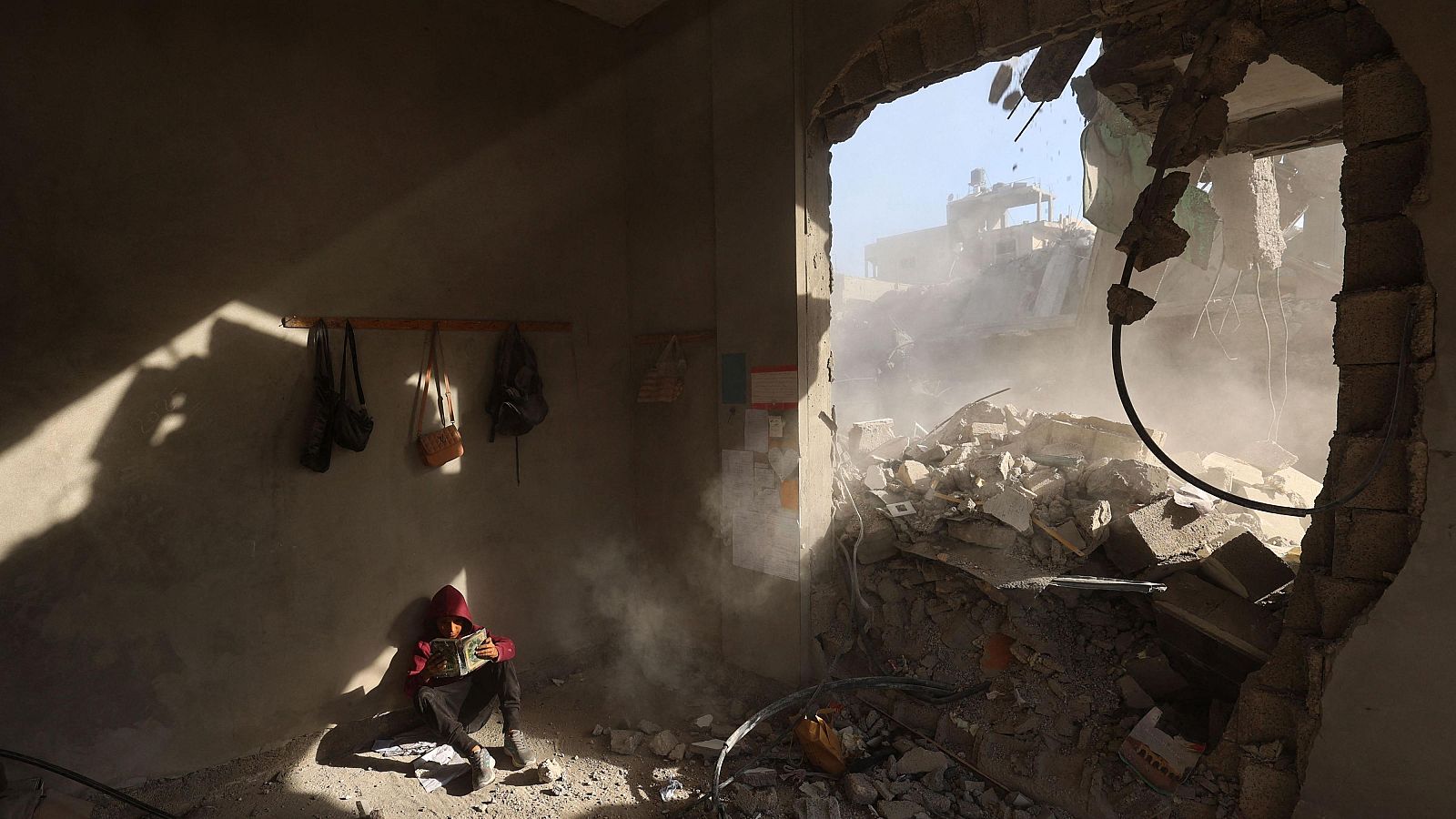 Un niño palestino sostiene un libro sentado entre los escombros de una casa