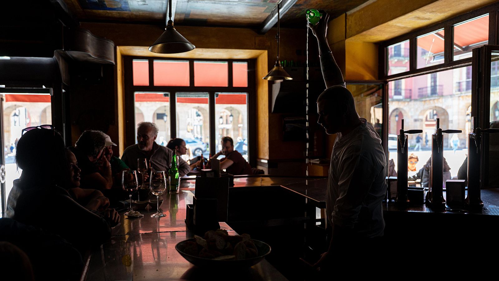 Silueta de camarero sirviendo sidra en un bar oscuro, clientes sentados en la barra durante un apagón. Ambiente contrastante.