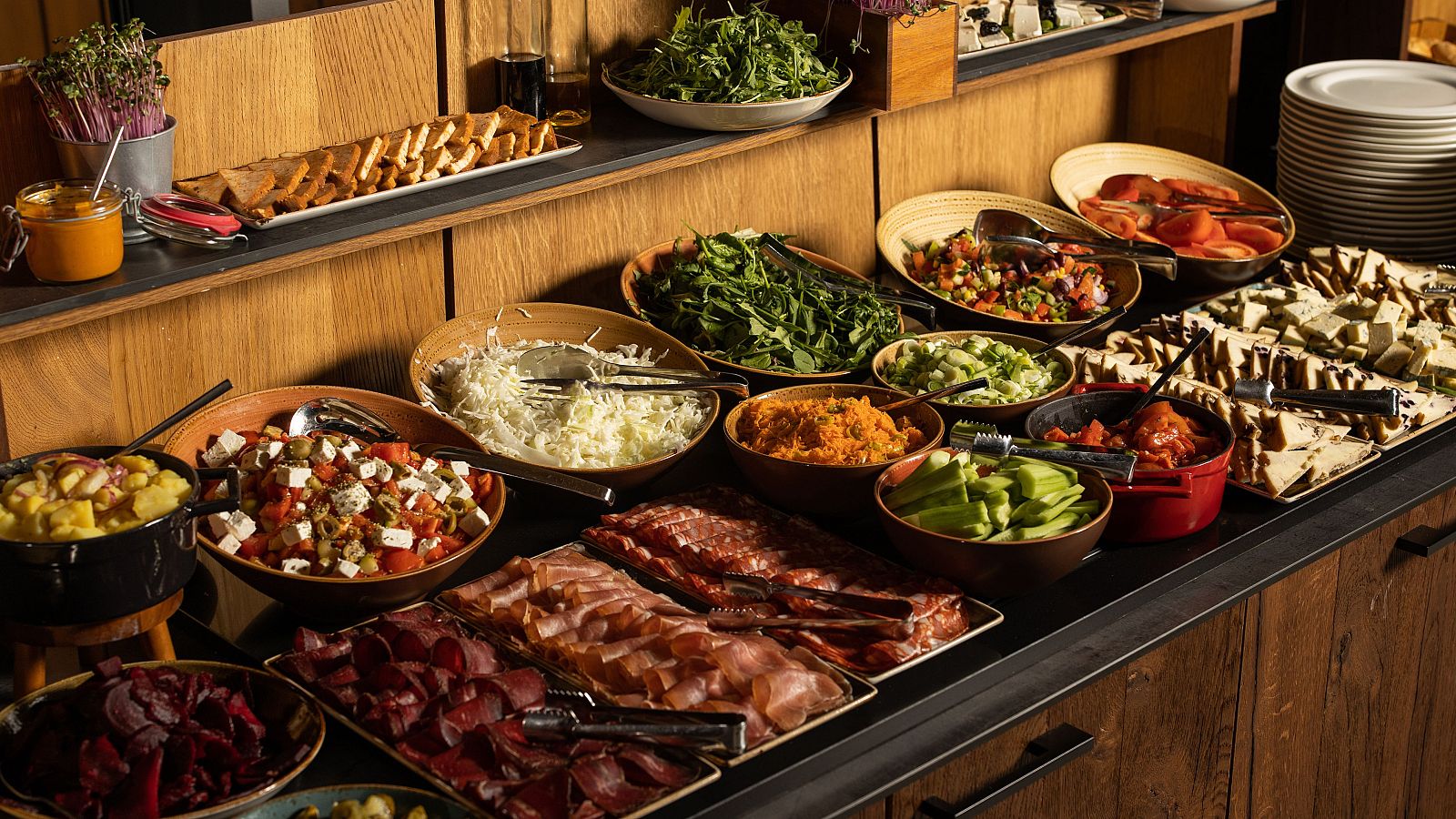 Buffet con amplia selección de comida: panes, ensaladas, guisos, embutidos y patatas. Recipientes de cerámica y madera sobre mesa oscura.